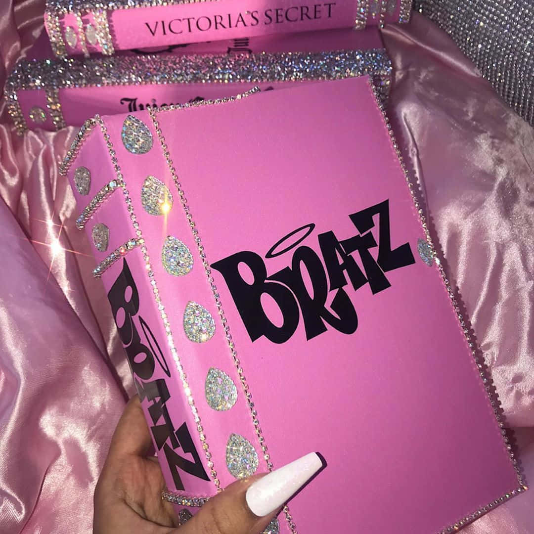 bratz 美學 桌布 壁紙 — 一隻手拿著一本閃亮的粉紅色Bratz書，帶有天使翅膀和水晶，背景是緞面粉紅色。