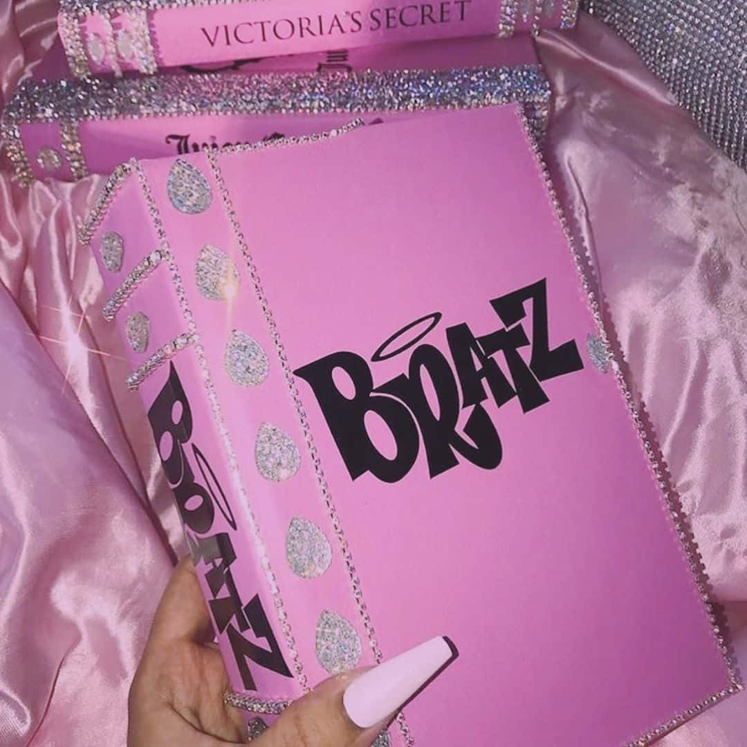 Pink Y2k 桌布 壁紙 — 一隻手拿著一本閃粉粉紅色Victoria's Secret 'Boatz'書，帶有銀色裝飾，背景是緞面粉紅色。