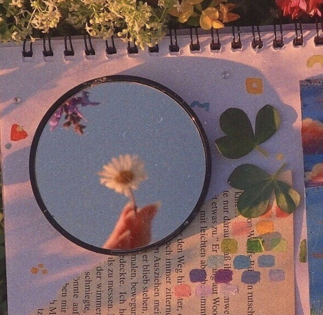 手拿雛菊倒映在鏡中，鏡子放在裝飾著三葉草和花朵的筆記本上。 — 田園風 GIF 壁紙