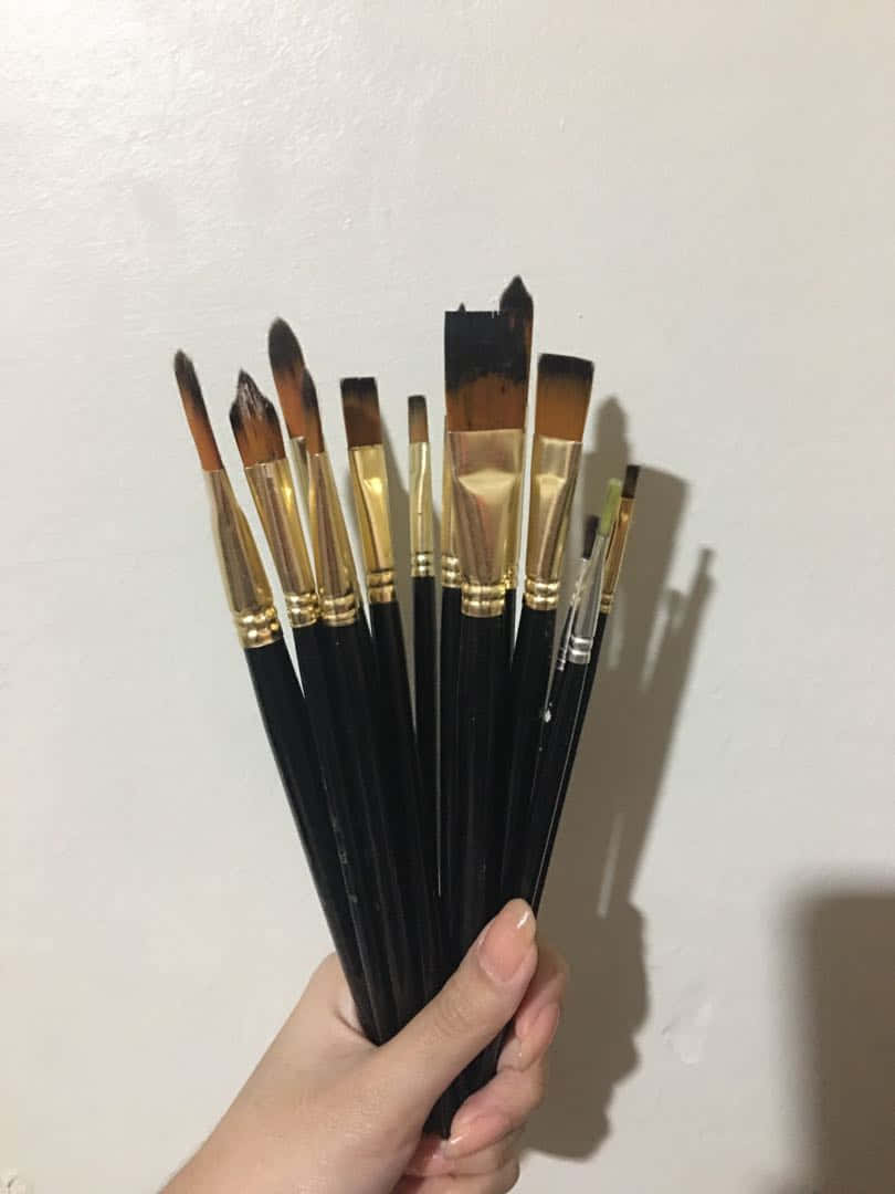 Paint Brush 桌布 壁紙 — 一隻手拿著一束用過的畫筆，黑色手柄和金色金屬箍，對著純白牆壁。