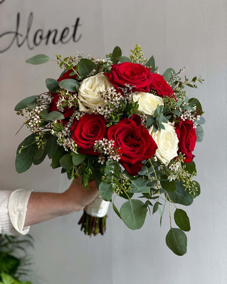 Roses Wedding 桌布 壁紙 — 一隻手拿著一束茂盛的紅白玫瑰，點綴著綠色桉樹和小白花，背景是素牆，上方寫有'Monet'。