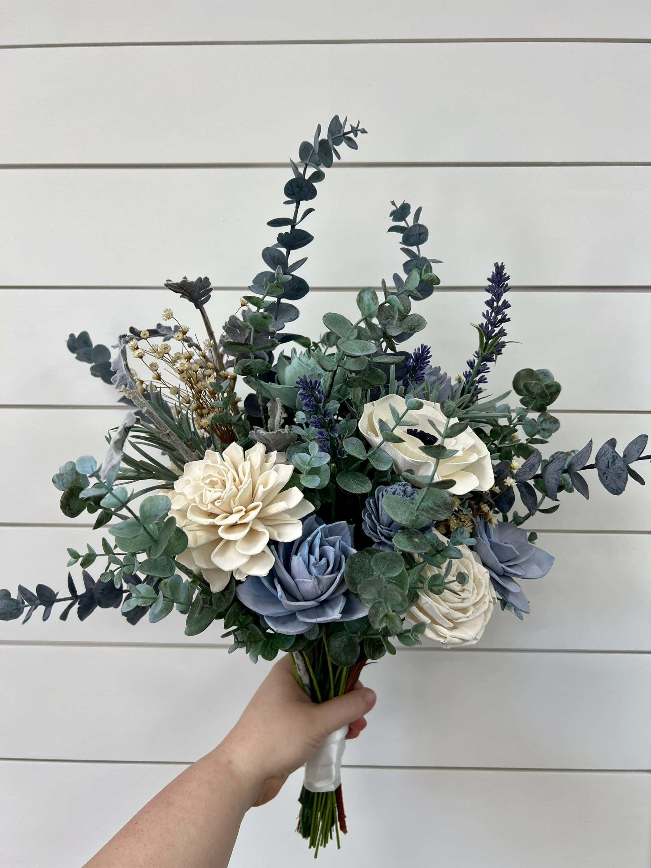 Bouquet 桌布 壁紙 — 一隻手拿著一束藍色和奶油色的茂盛花束，搭配尤加利葉，背景是白色的船型板牆。