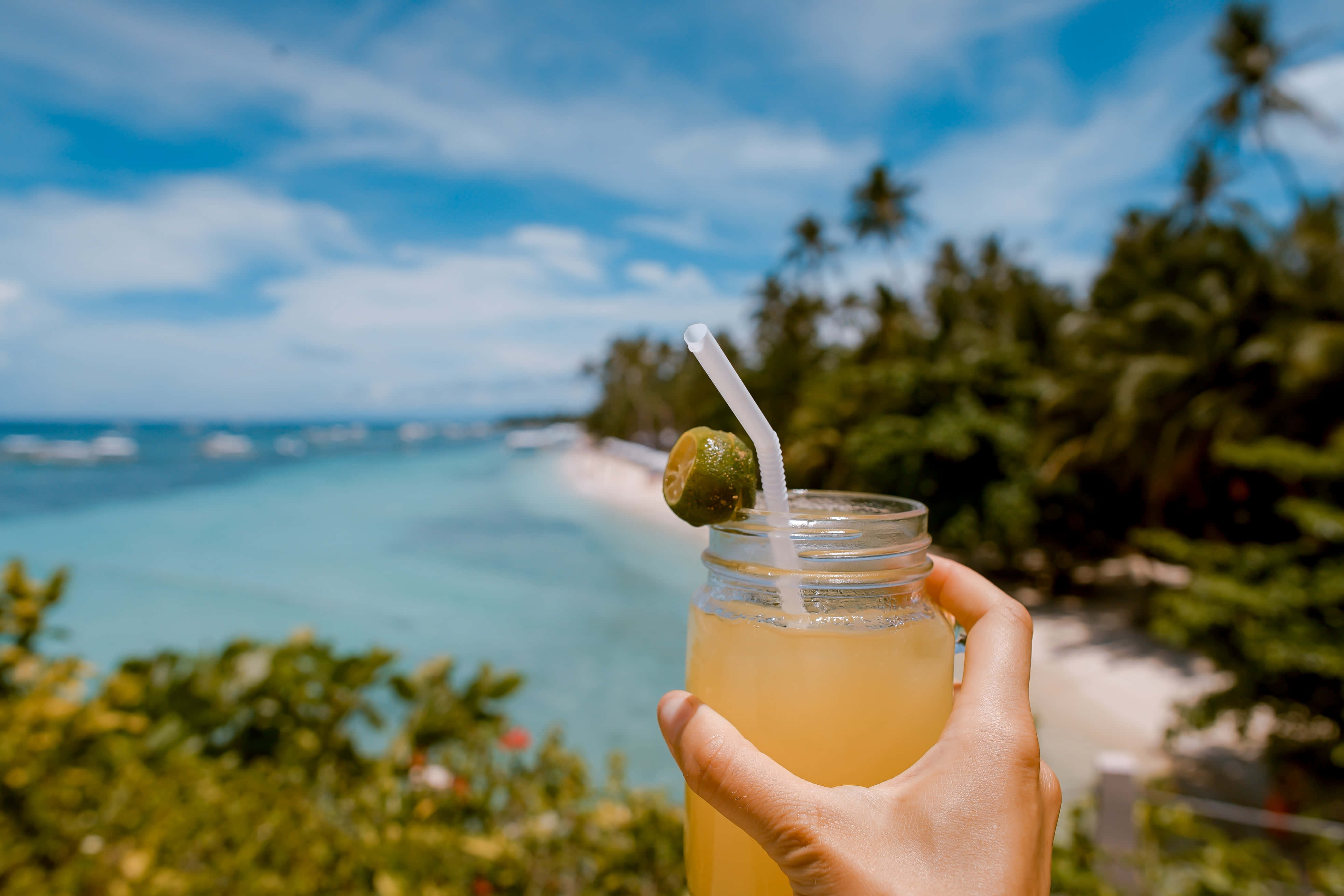 Summer Vacation 桌布 壁紙 — 一隻手拿著一杯有青檸和吸管的熱帶飲品，背景是模糊的藍綠色海洋和棕櫚樹，上方是明亮的藍天。