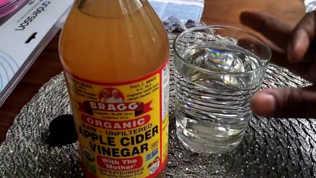 Drink 桌布 壁紙 — 一隻手拿著一杯水，旁邊放著一瓶Bragg有機蘋果醋，放在銀色紋理墊上。