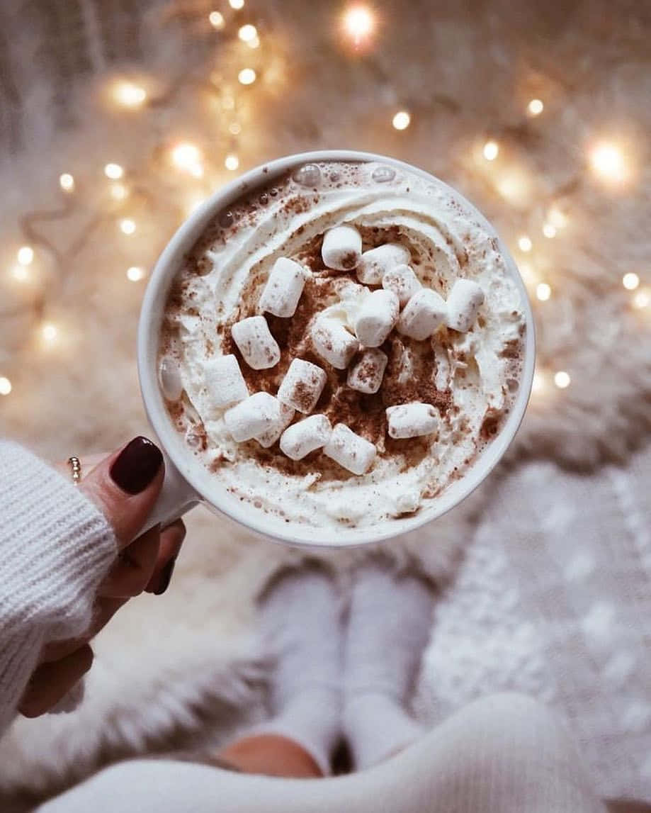 Hot Chocolate 桌布 壁紙 — 一隻手拿著一杯熱巧克力，上面有鮮奶油和迷你棉花糖，背景是溫馨的閃爍小燈。