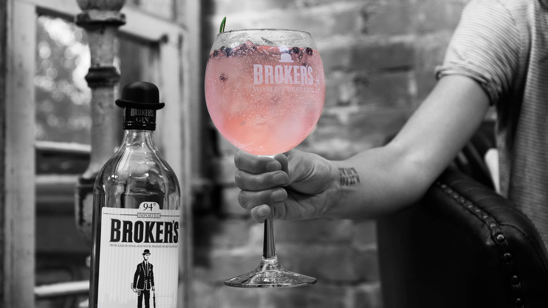 Drink 桌布 壁紙 — 一隻手拿著一杯粉紅色琴酒雞尾酒，旁邊有一瓶Brokers琴酒，背景是磚牆，帶有選擇性色彩。