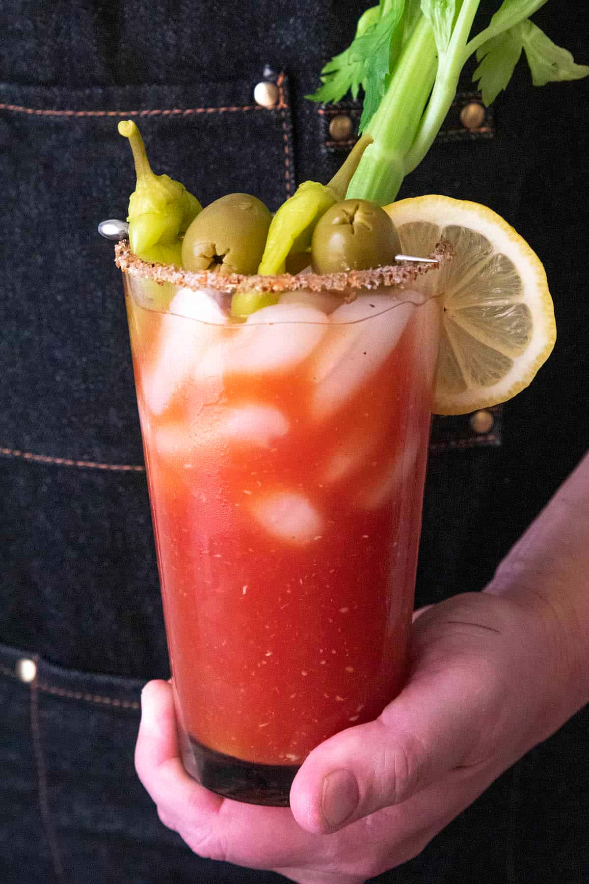 Beautiful Drink 桌布 壁紙 — 一隻手拿著一杯鮮紅色的血腥瑪麗，飾以橄欖、青椒、芹菜和檸檬片，背景為牛仔布。