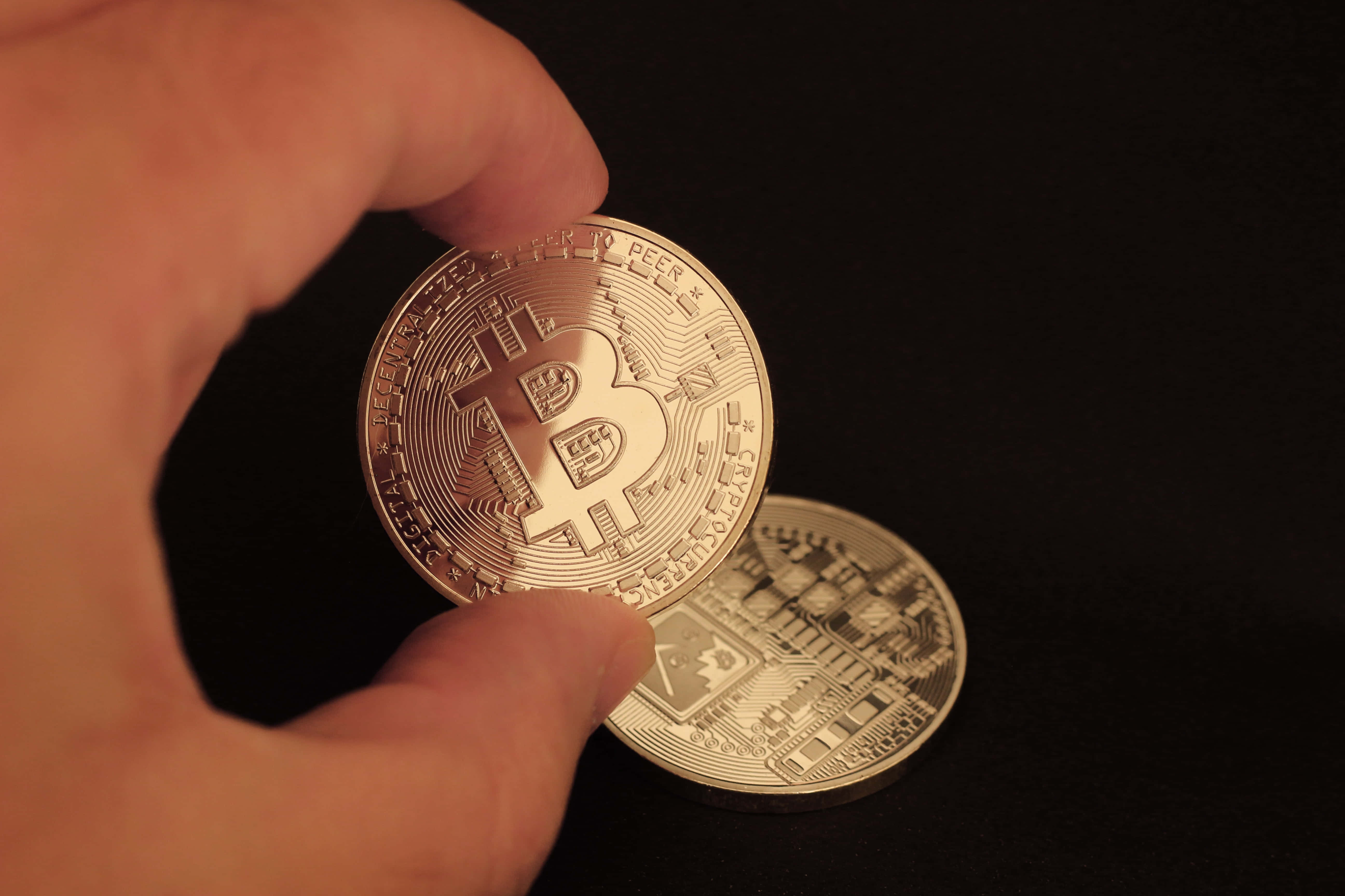 Mining 桌布 壁紙 — 一隻手拿著一枚閃閃發光的金色比特幣，背景是純黑色，下方還有另一枚硬幣。