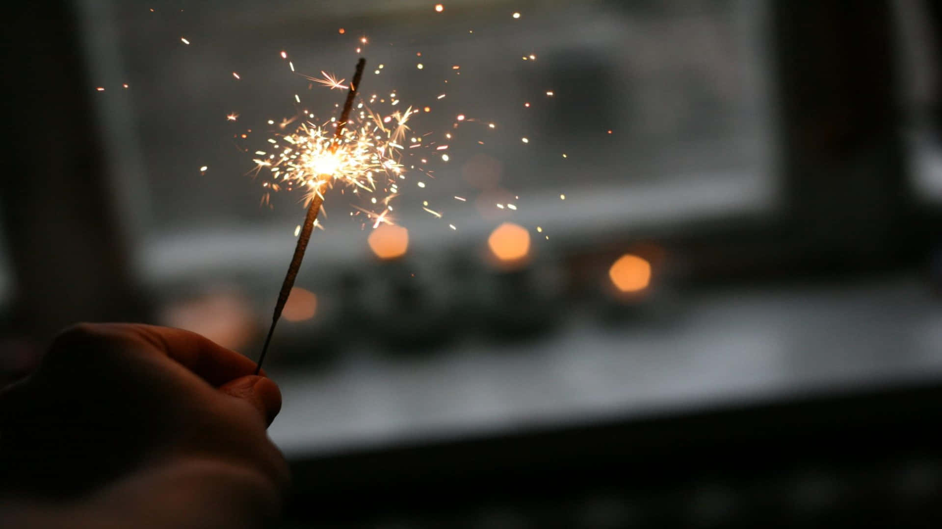 節日 桌布 壁紙 — 一隻手拿著一根閃爍的煙火棒，在黑暗模糊的背景中發出金色火花。