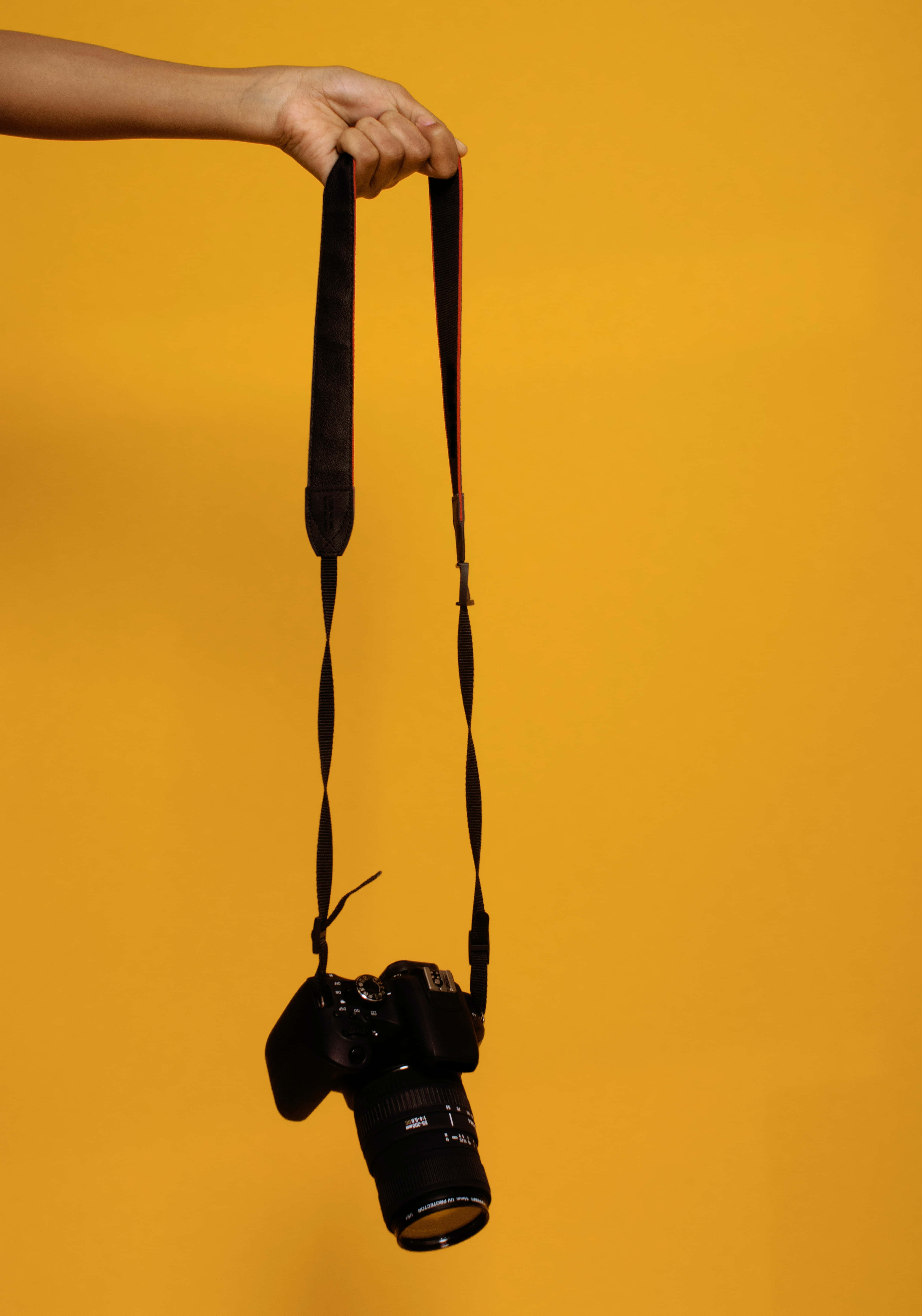Dslr 桌布 壁紙 — 一隻手拿著一條黑色單反相機的帶子，背景是純暖黃色。