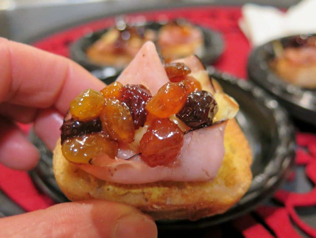 Black Forest Ham 桌布 壁紙 — 一隻手拿著一款美食克羅斯蒂尼，上面放有閃亮的蜜餞水果和火腿，襯托著節日紅色桌布。