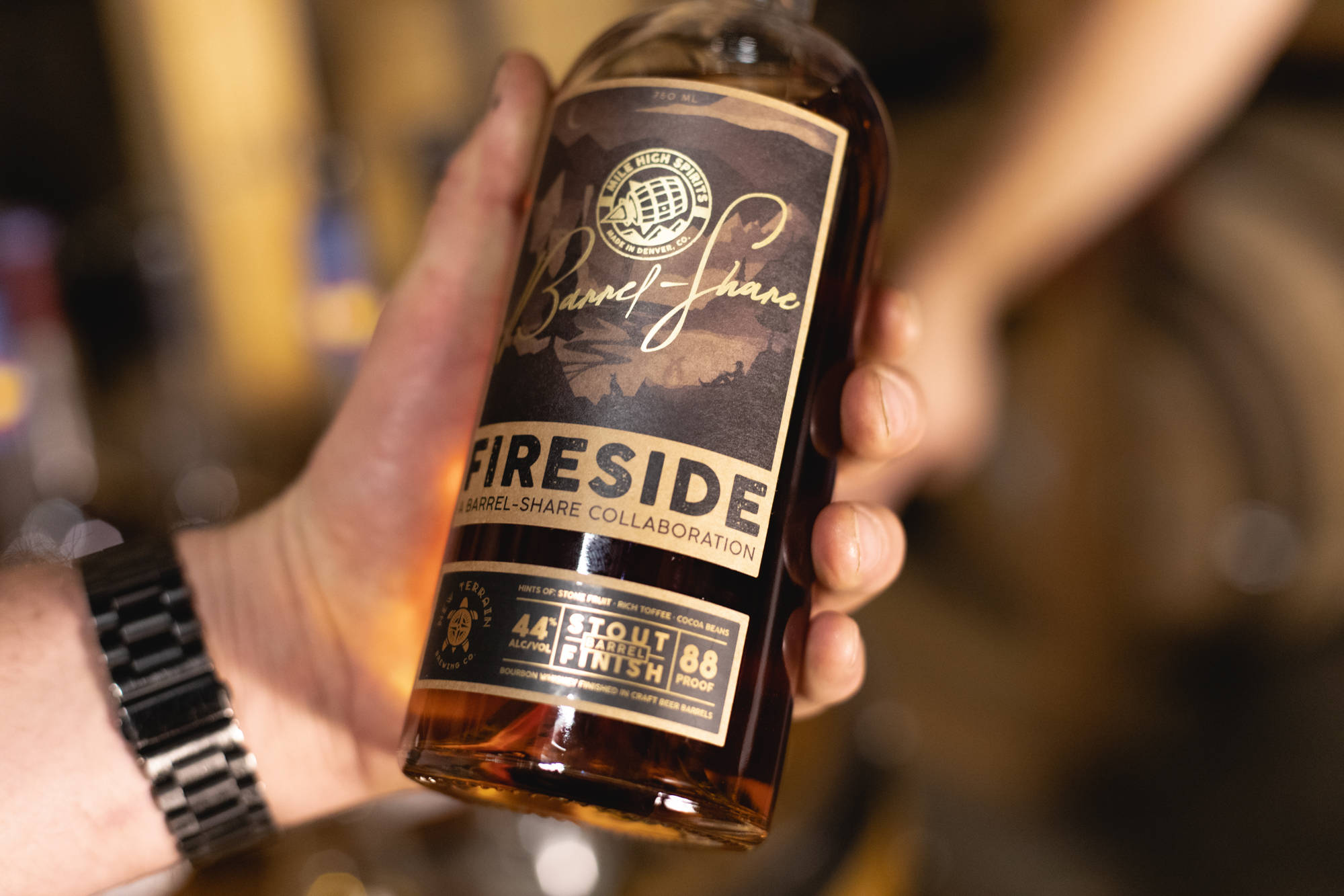 Blur 桌布 壁紙 — 一隻手拿著一瓶 Fireside Barrel-Share Collaboration 黑啤酒威士忌，琥珀色液體豐富，標籤精細，背景是溫暖模糊的酒吧。