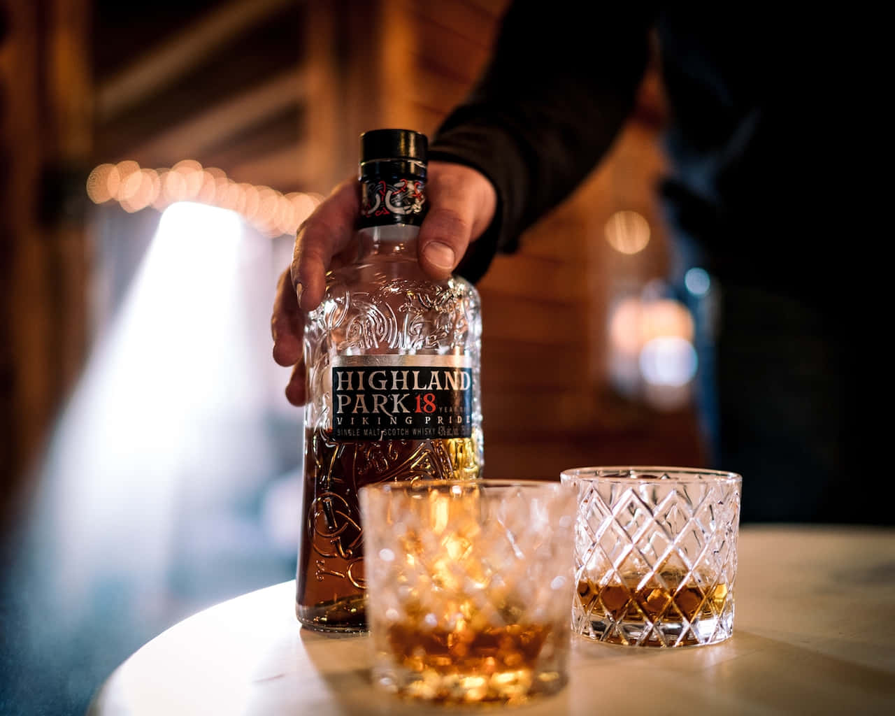 glasses 桌布 壁紙 — 一隻手拿著一瓶Highland Park 18 Year Old Viking Pride威士忌，旁邊有兩個裝滿琥珀色液體的水晶杯放在木桌上。