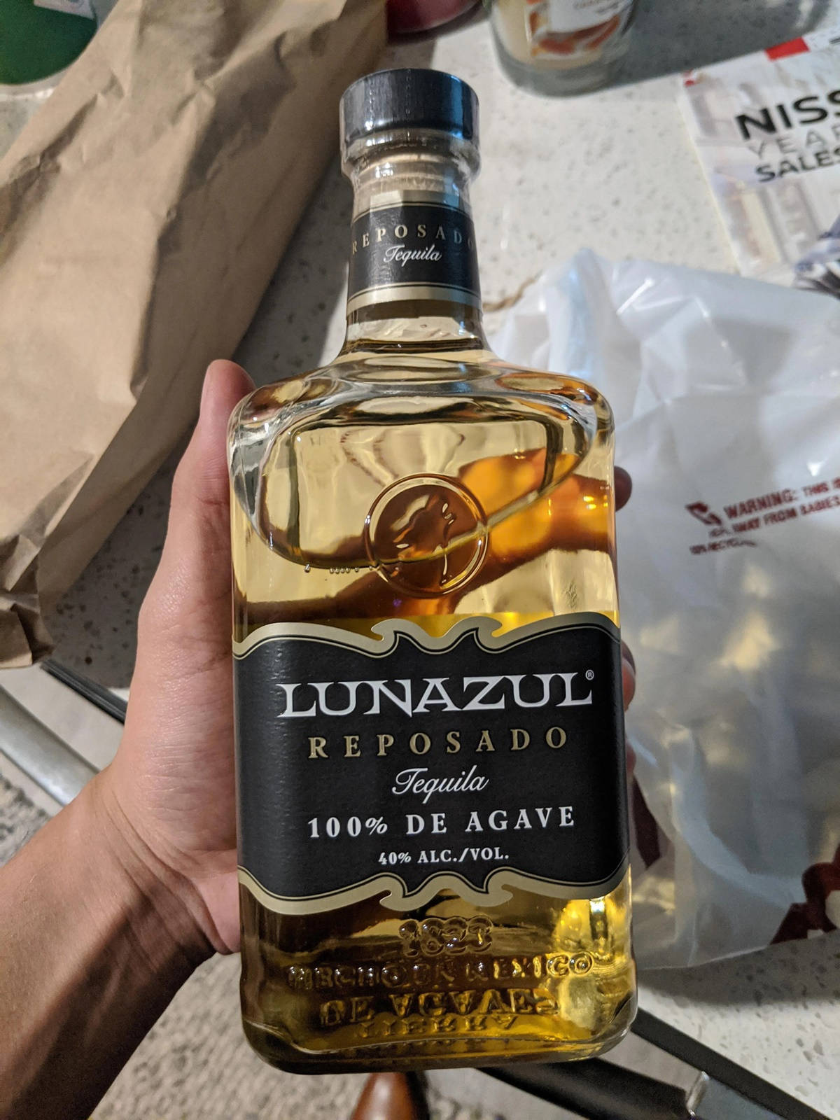 alcohol 桌布 壁紙 — 一隻手拿著一瓶Lunazul Reposado龍舌蘭酒，金色液體透過透明玻璃清晰可見，背景是雜亂的檯面。