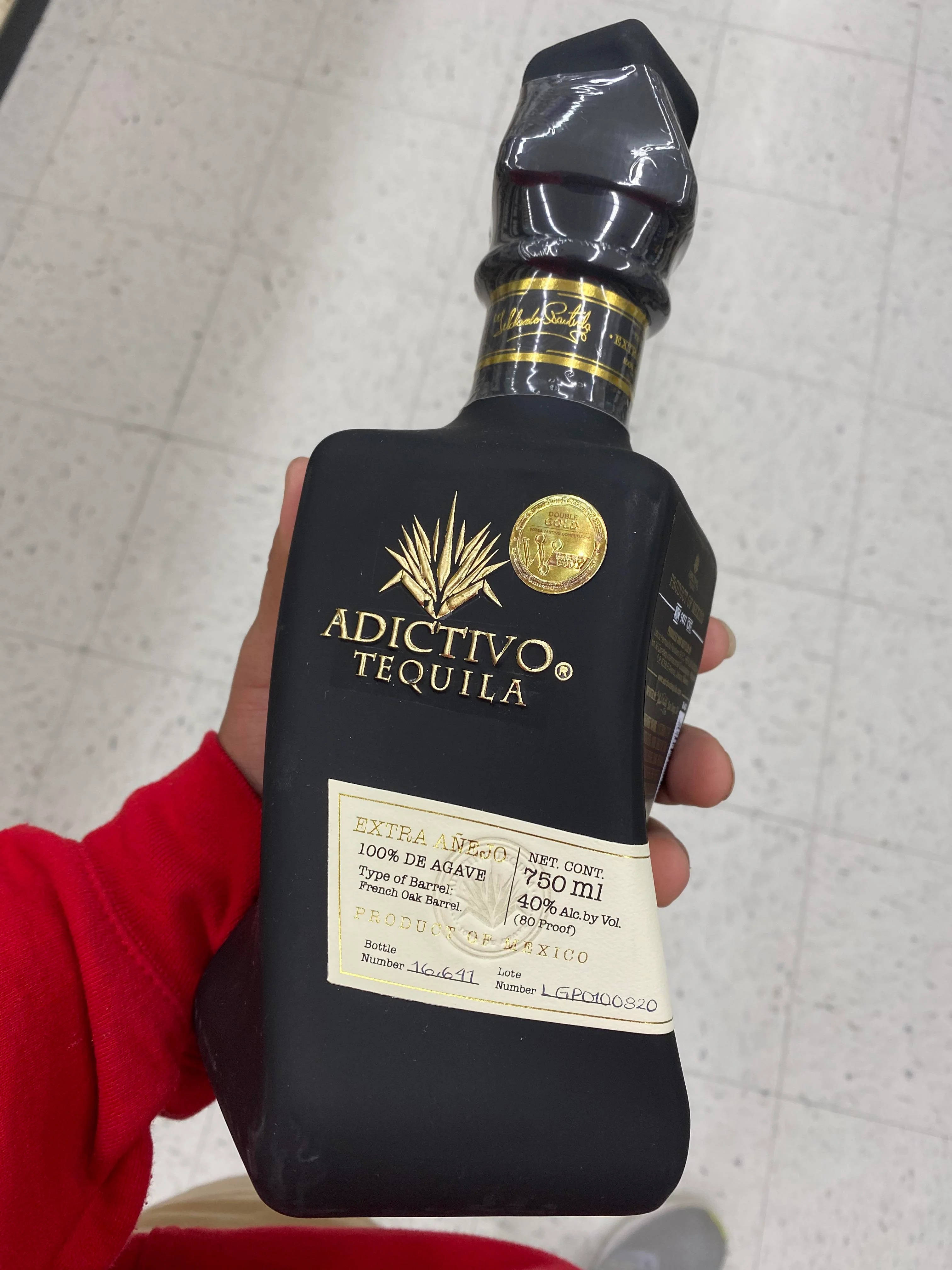 iPhone 桌布 壁紙 — 一隻手拿著一瓶帶有金色裝飾的時尚黑色Adictivo Tequila酒瓶，背景是瓷磚地板。