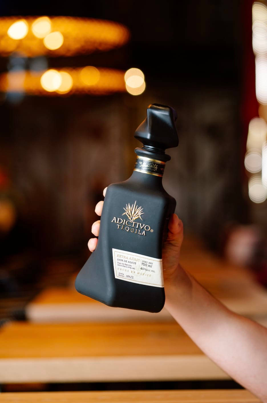 手機 桌布 壁紙 — 一隻手拿著一瓶時尚的黑色Adictivo Tequila，背景是溫暖燈光的酒吧，帶有模糊的金色散景燈光。