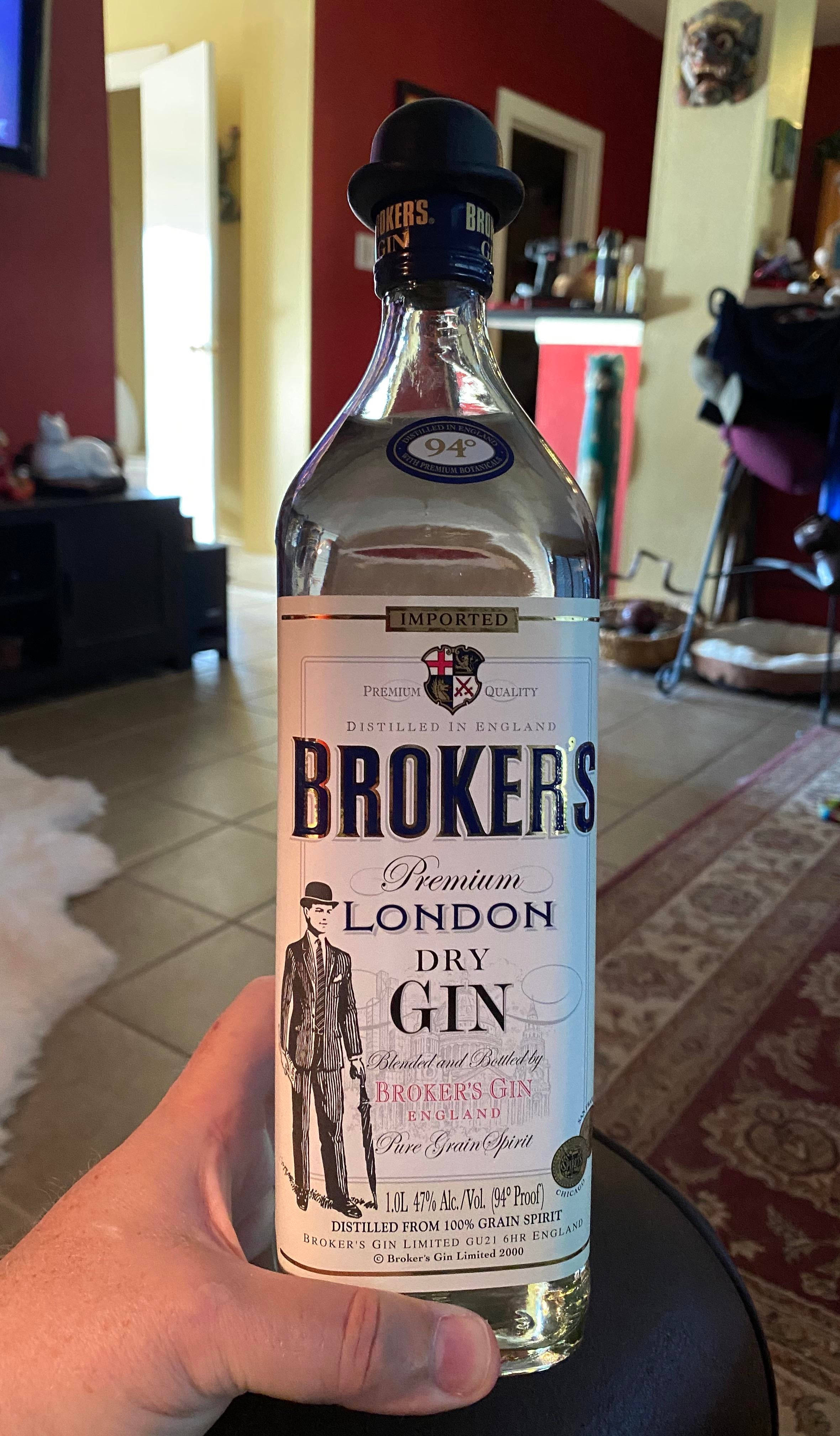 bottle 桌布 壁紙 — 一隻手拿著一瓶清澈的Brokers London Dry Gin，瓶身帶有復古標籤，上面印有一位身穿西裝、戴著高帽的男士，背景是溫暖燈光的家居內飾。