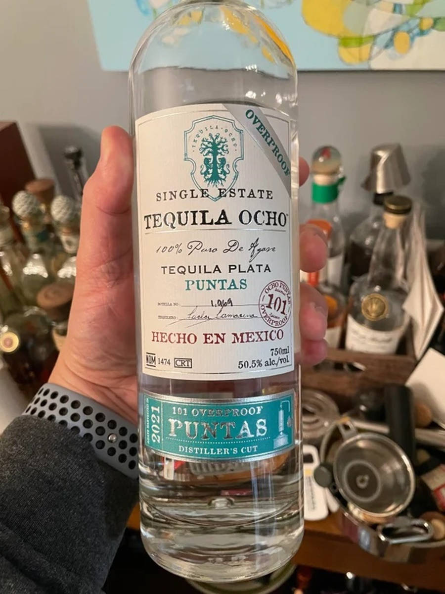 bottle 桌布 壁紙 — 一隻手拿著一瓶透明的101 proof Tequila Ocho，背景是酒瓶和酒吧工具。
