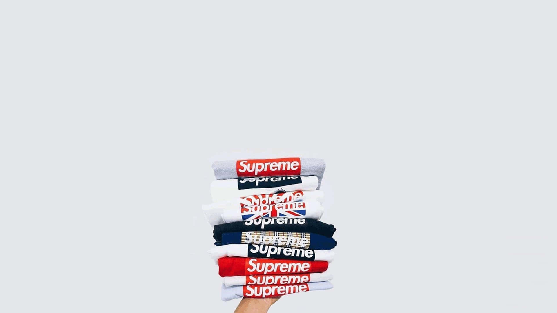 supreme aesthetic 桌布 壁紙 — 一隻手拿著一疊折疊好的Supreme品牌T恤，背景是純淺灰色。
