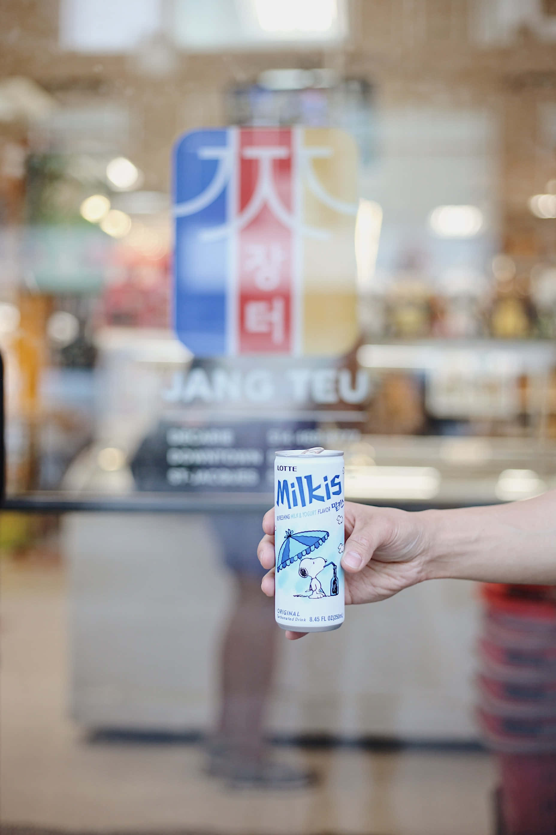 手機 桌布 壁紙 — 一隻手拿著一罐白色的Lotte Milkis，罐上有史努比在雨傘下，背景是模糊的商店。