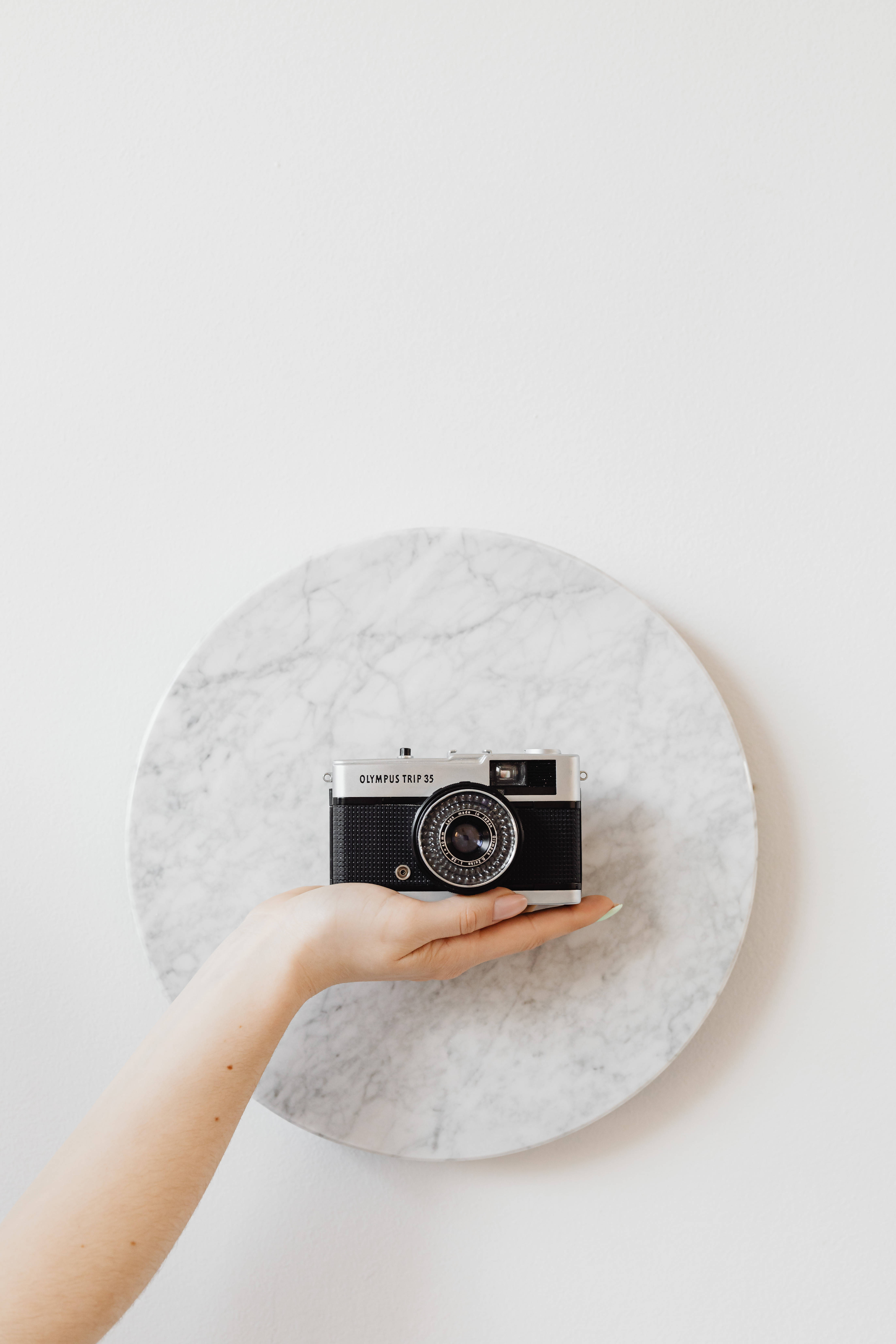 相機 桌布 壁紙 — 一隻手拿著一部復古 Olympus Trip 35 相機，放在白色大理石盤中央，背景是素色牆壁。
