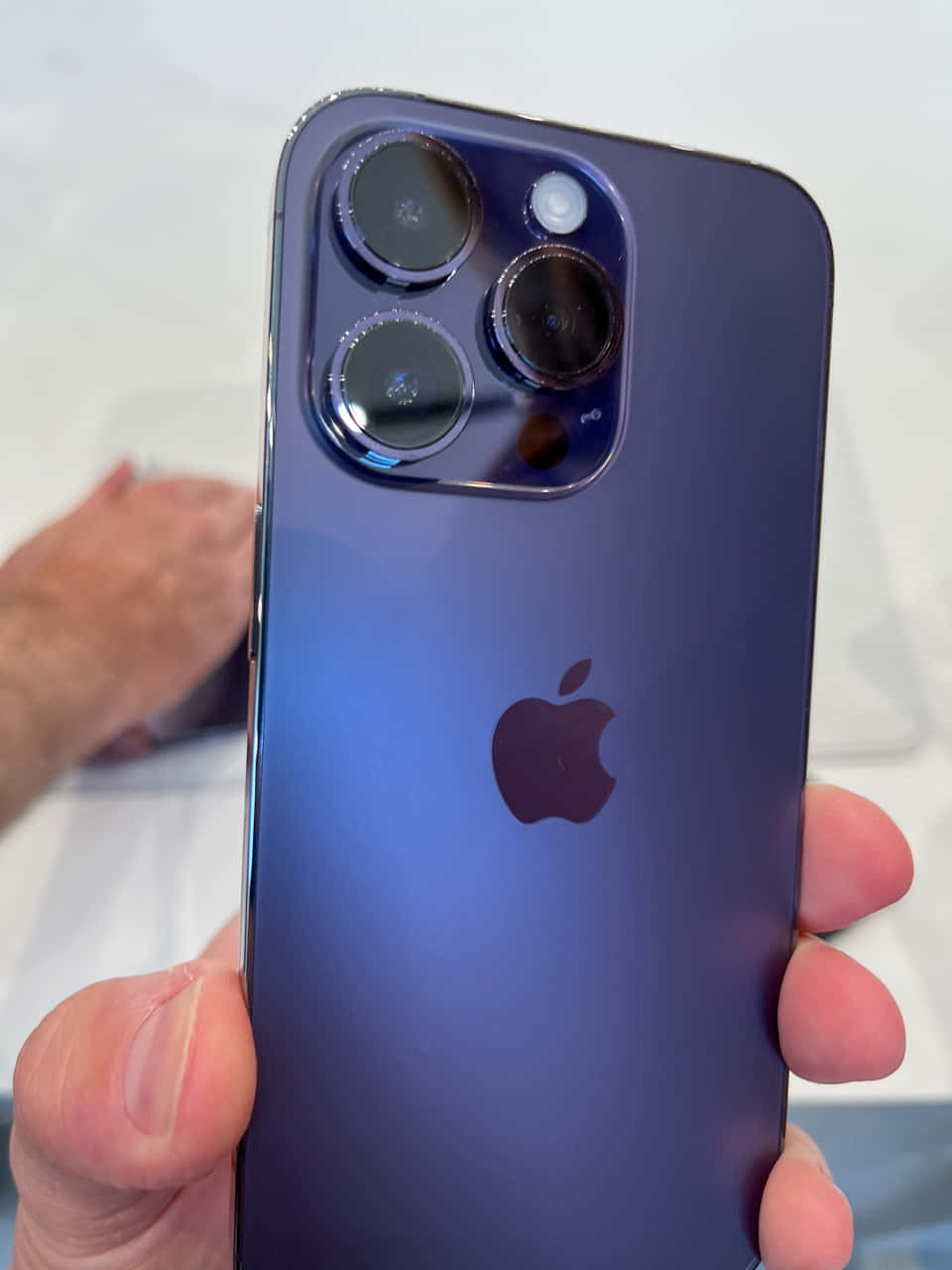 iPhone 桌布 壁紙 — 一隻手拿著一部時尚的紫色iPhone，配有突出的三鏡頭系統和Apple標誌。