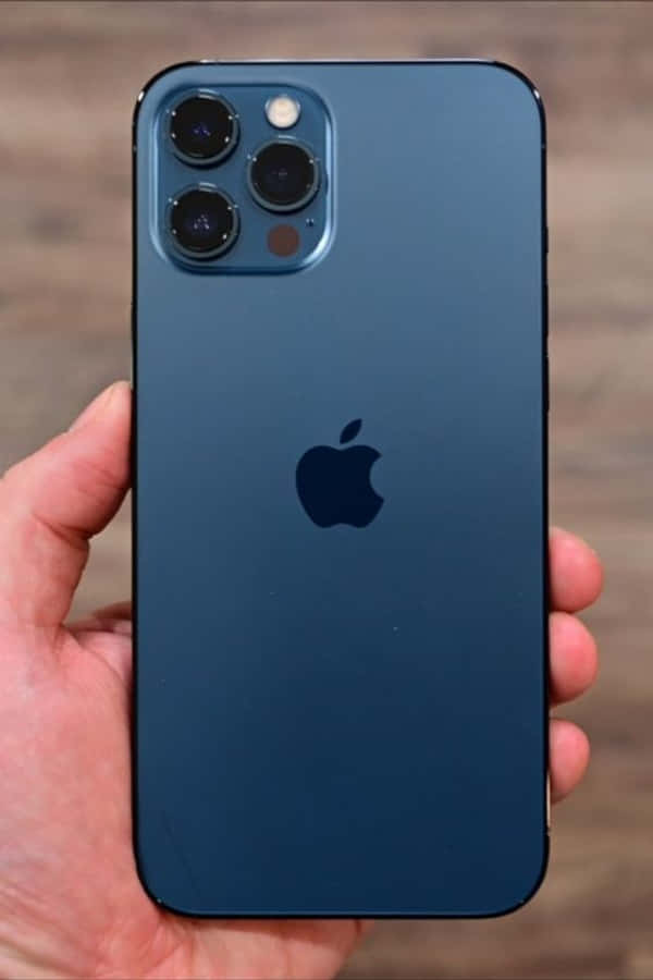 iPhone 12 桌布 壁紙 — 一隻手拿著一部時尚的藍色iPhone，帶有突出的相機模組和Apple標誌，背景是模糊的木質背景。