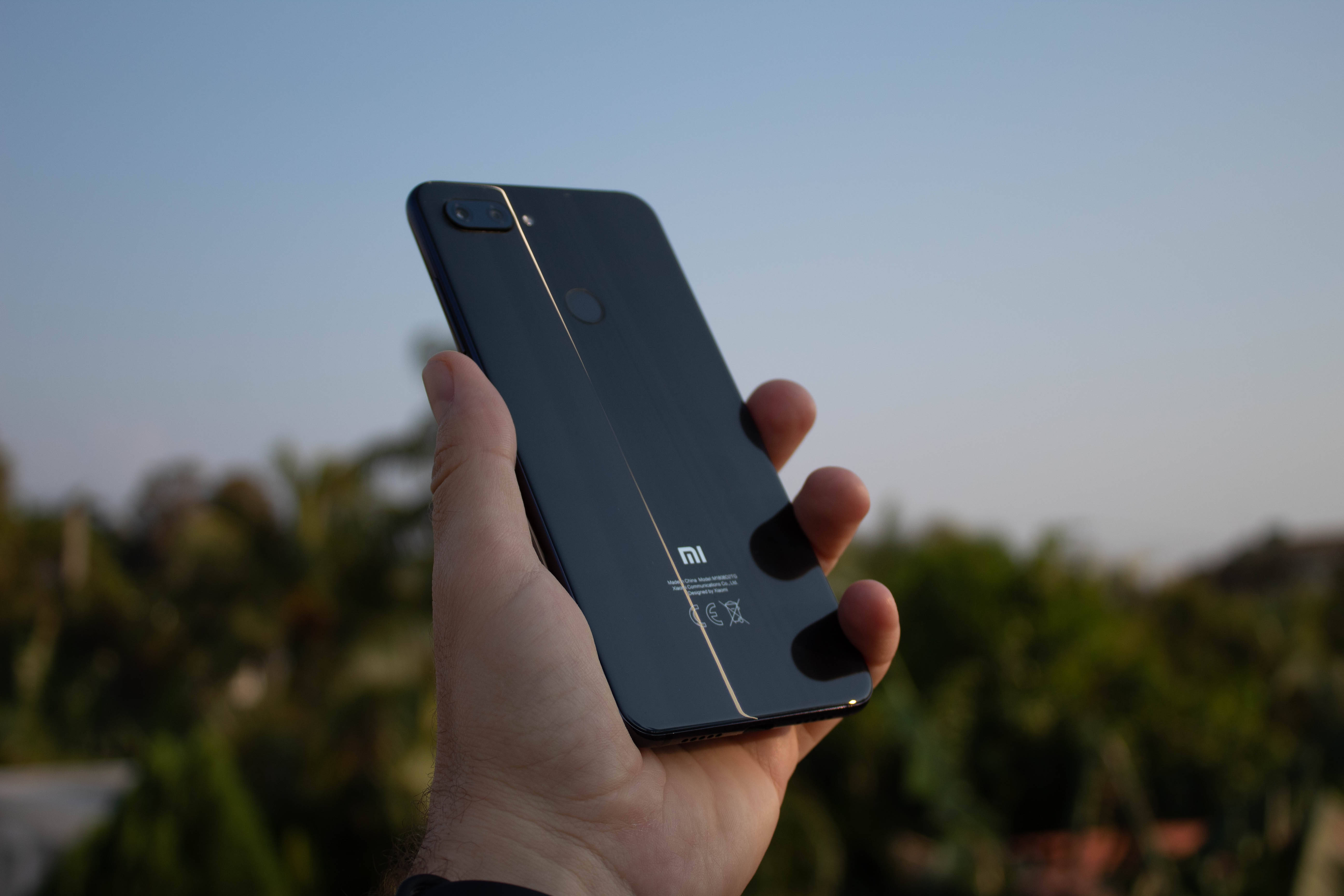 Xiaomi 桌布 壁紙 — 一隻手拿著一部時尚的黑色Xiaomi智慧型手機，背景是柔和的戶外環境，帶有溫暖的傍晚光線。