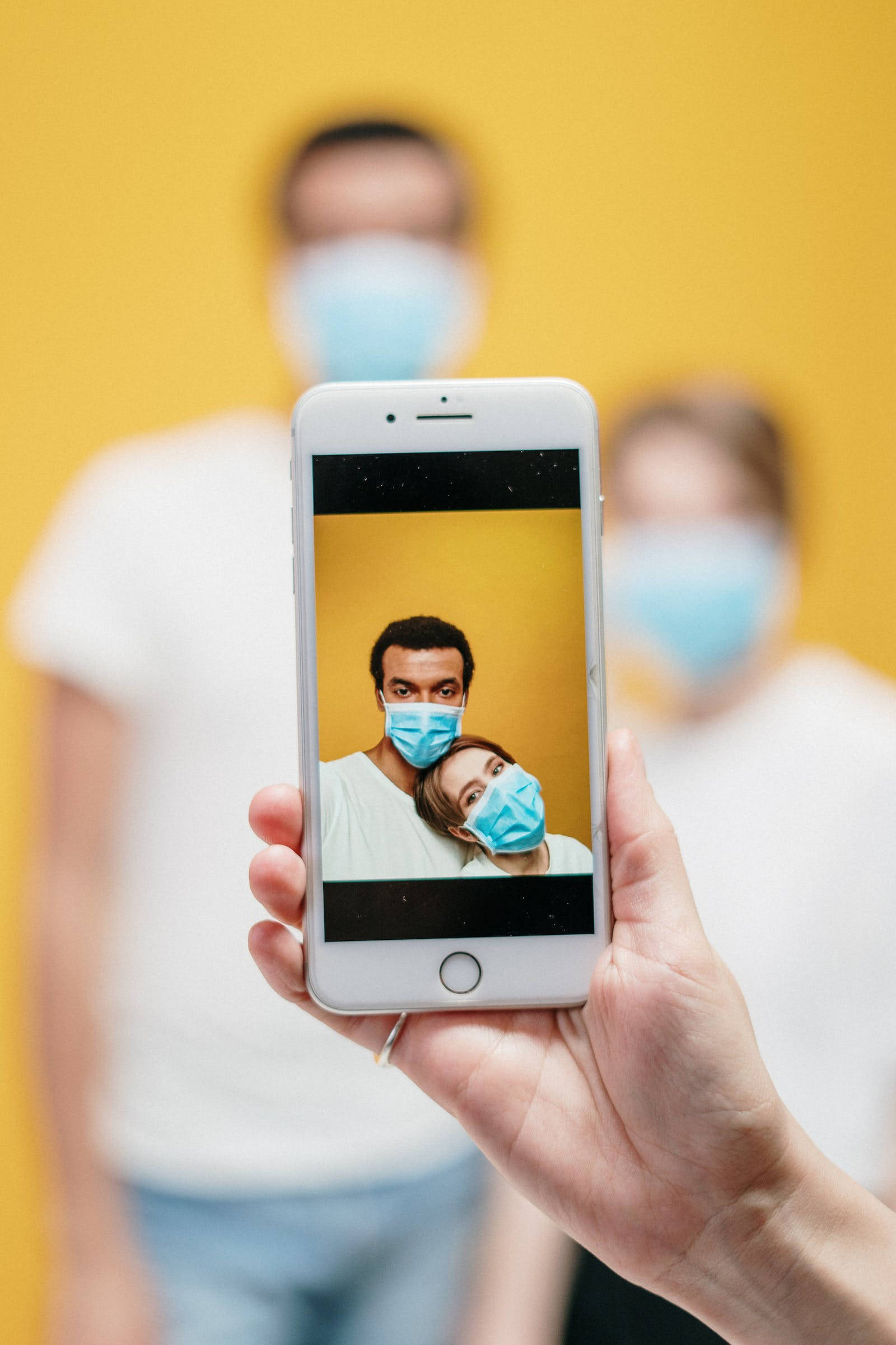 Love iPhone 桌布 壁紙 — 一隻手拿著一部智慧型手機，顯示一對情侶戴著藍色口罩的自拍照，背景為鮮黃色。