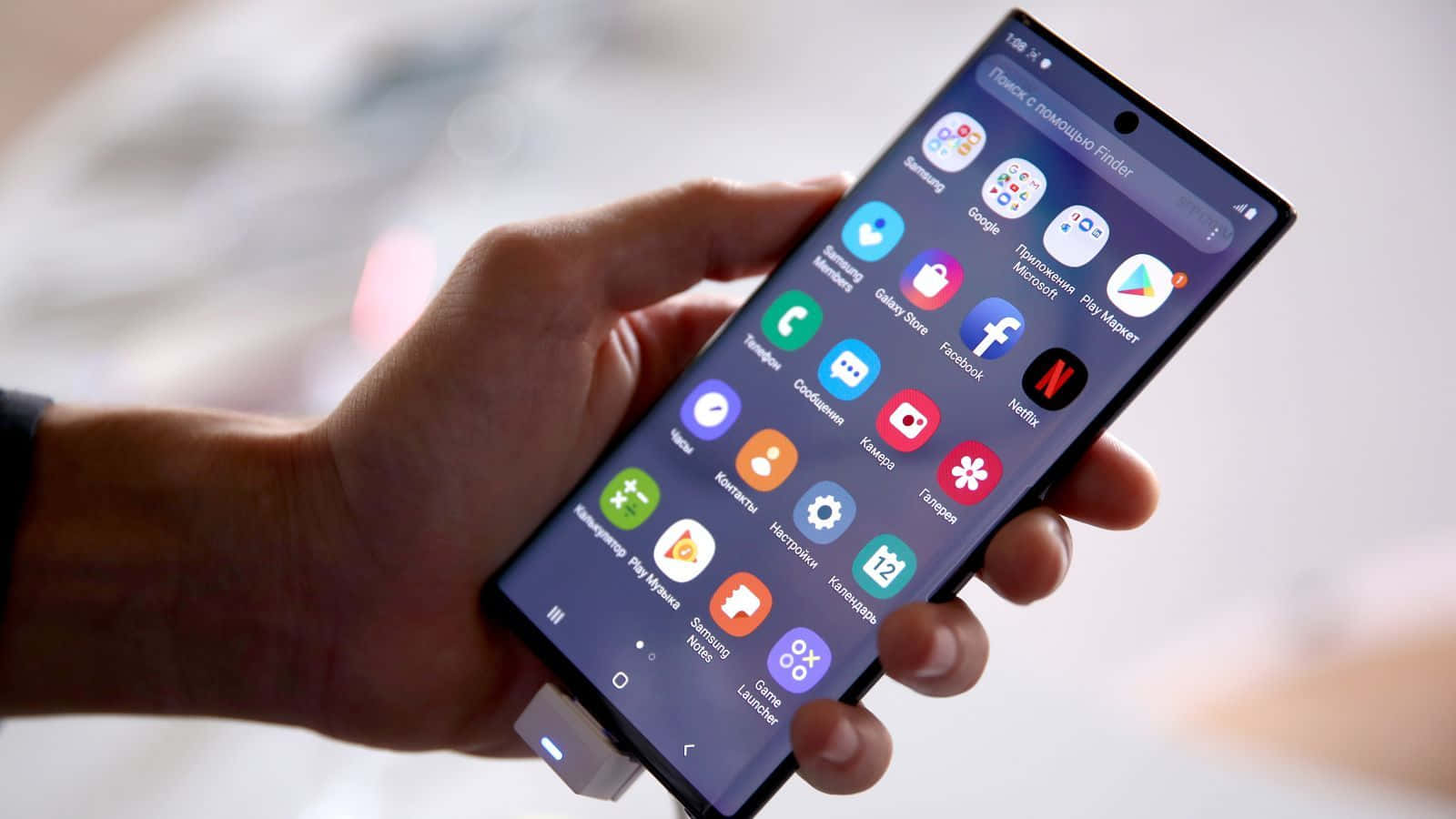 Note 10 桌布 壁紙 — 一隻手拿著一部現代智慧型手機，螢幕上顯示一個色彩繽紛的應用程式網格，帶有俄語標籤，背景柔和模糊。