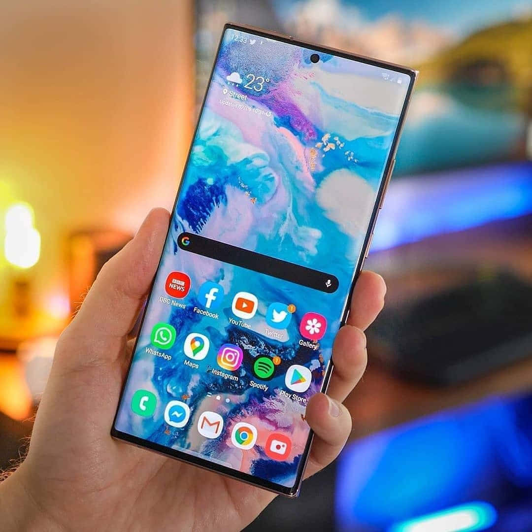 Note 10 桌布 壁紙 — 一隻手拿著一部現代智慧型手機，顯示出充滿活力的抽象桌布和應用程式圖示，背景是模糊的溫暖室內環境。