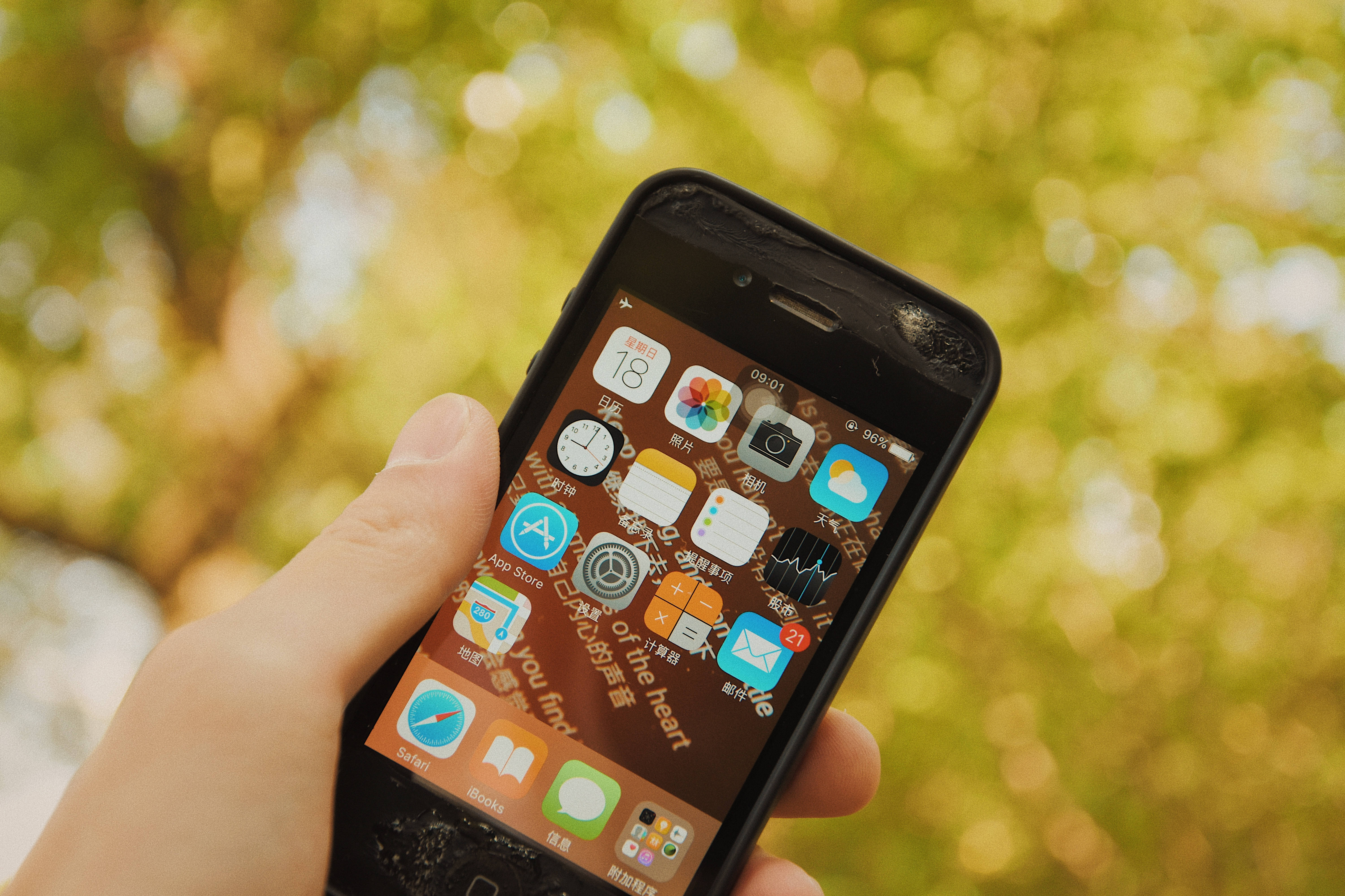 iOS 桌布 壁紙 — 一隻手拿著一部破裂的智慧型手機，顯示色彩繽紛的應用程式網格，背景是柔和模糊的綠色樹葉。
