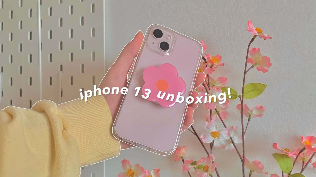可愛 iPhone 桌布 桌布 壁紙 — 一隻手拿著一部粉紅色iPhone 13，上面有一個花朵彈出式插座，背景是粉彩花朵和穿孔牆壁。