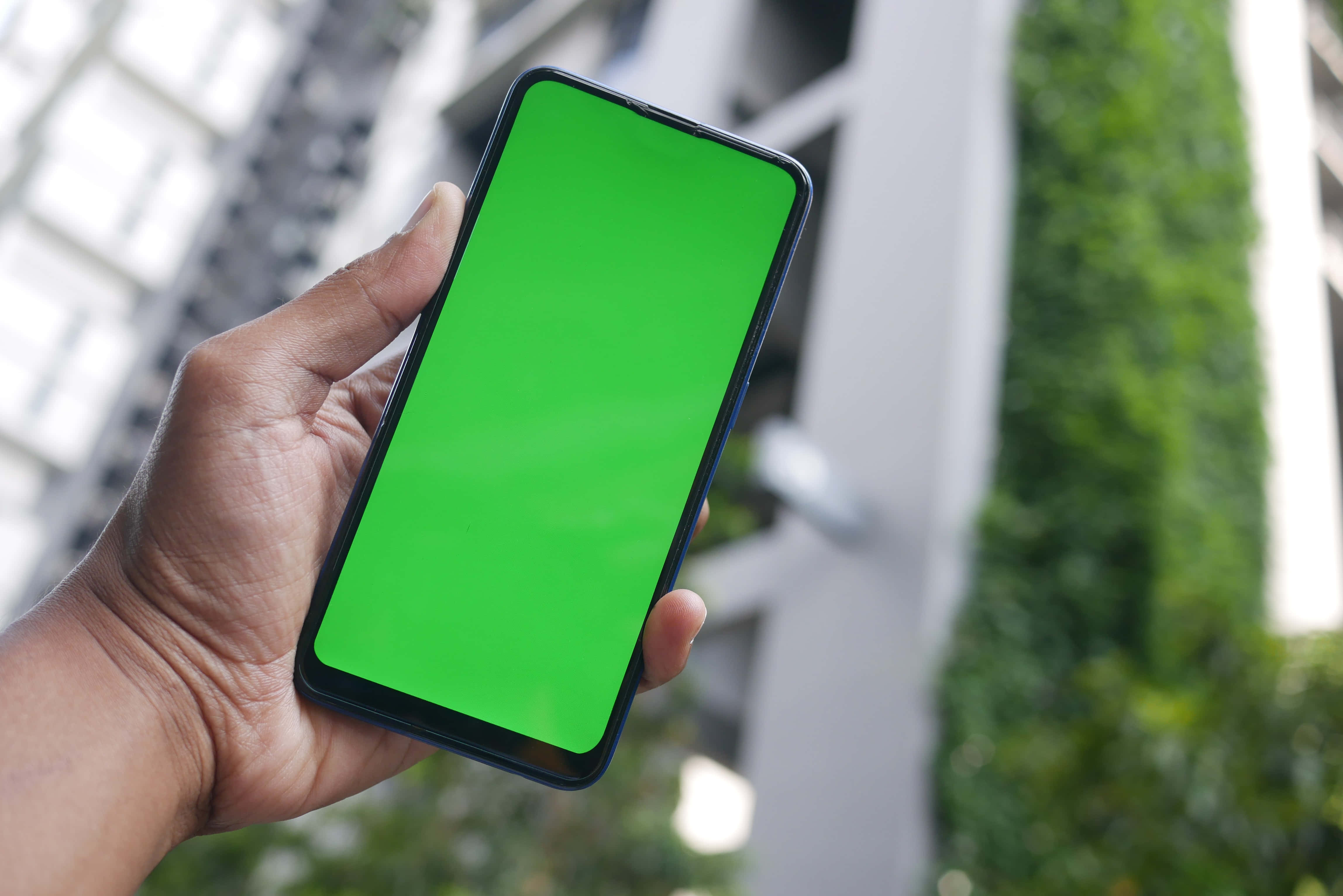 Smartphone 桌布 壁紙 — 一隻手拿著一部螢幕亮綠色的智慧型手機，背景是模糊的城市景觀和綠化。