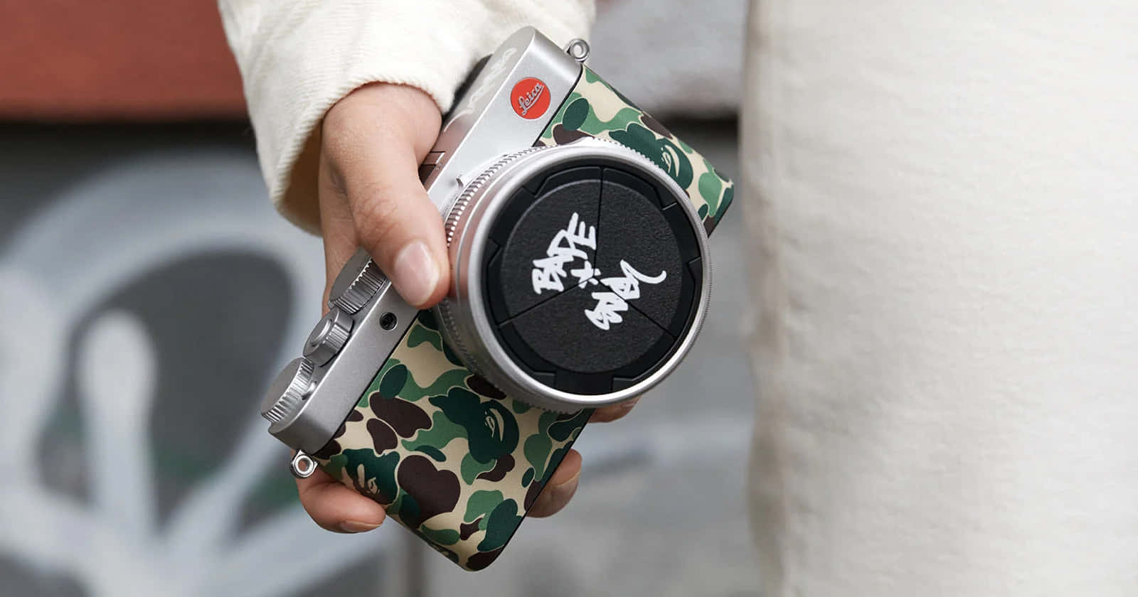 Bape 動態 桌布 壁紙 — 一隻手拿著一部銀色 Leica 相機，配有迷彩圖案機殼和帶有白色塗鴉風格文字的鏡頭蓋。