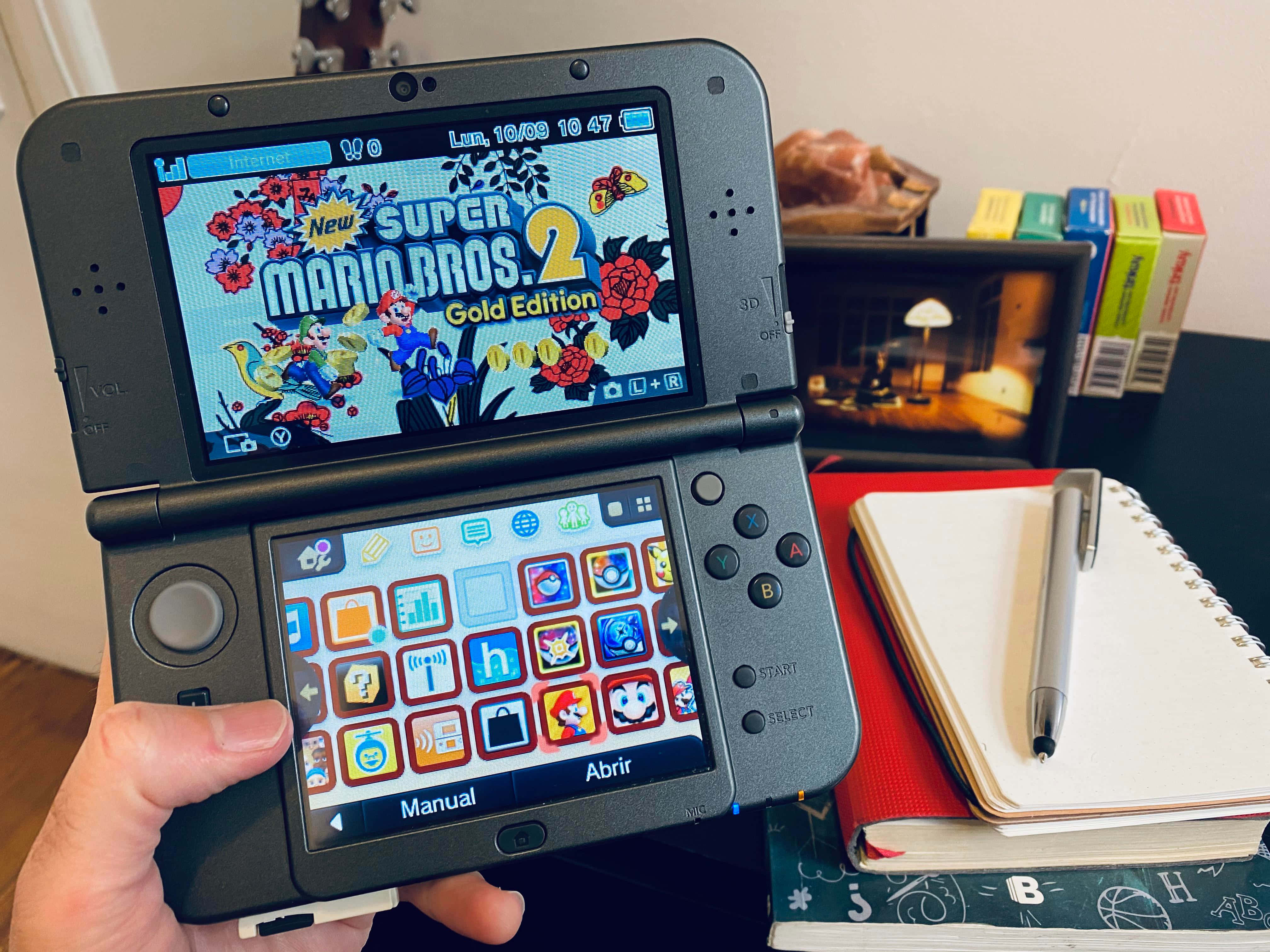 Casual Games 桌布 壁紙 — 一隻手拿著一部顯示New Super Mario Bros 2 Gold Edition的Nintendo 3DS，背景有筆記本筆和書籍。