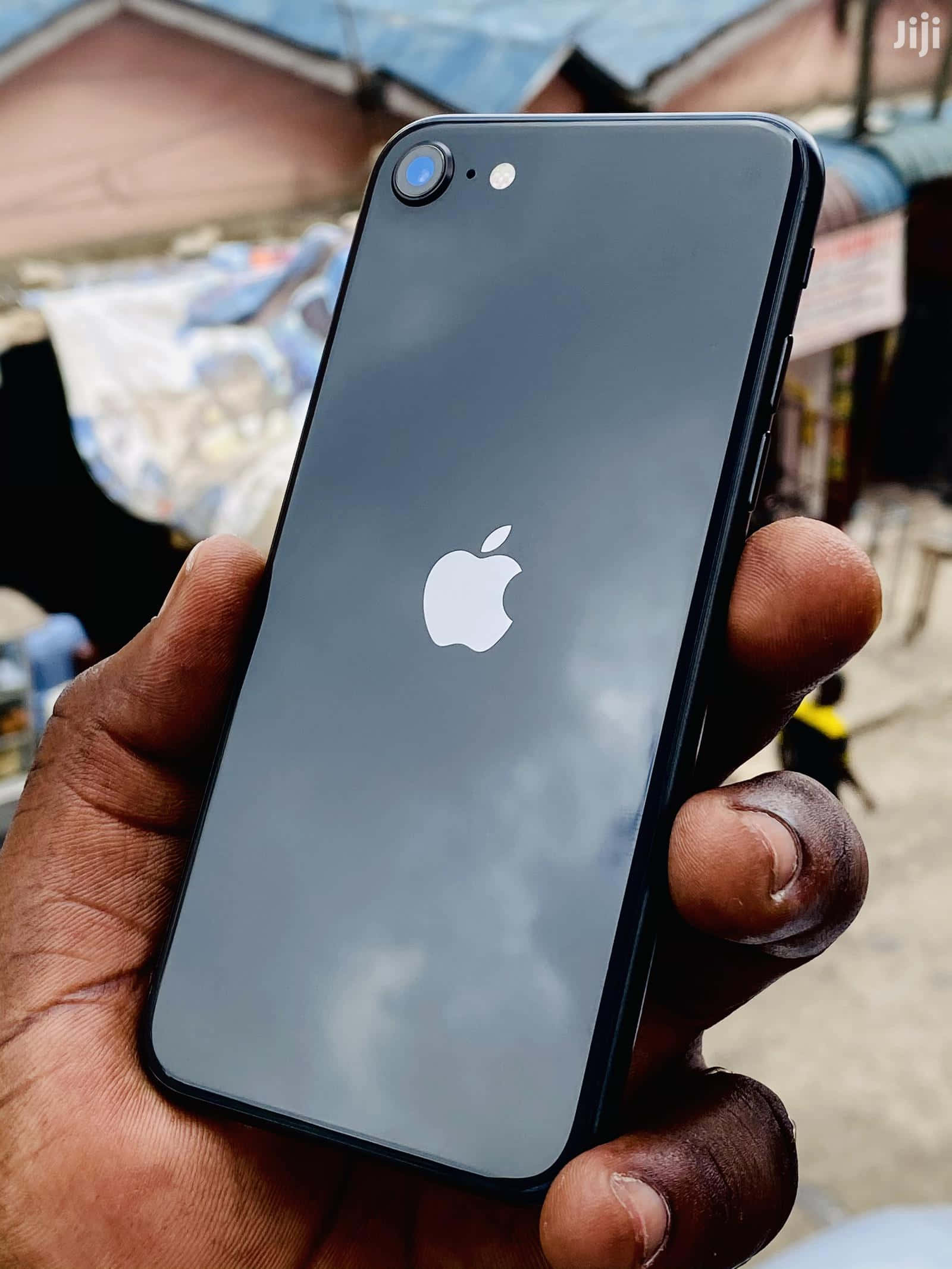 Apple Phone 桌布 壁紙 — 一隻手拿著一部黑色iPhone，背面反光顯示多雲的天空，背景是模糊的戶外市場。