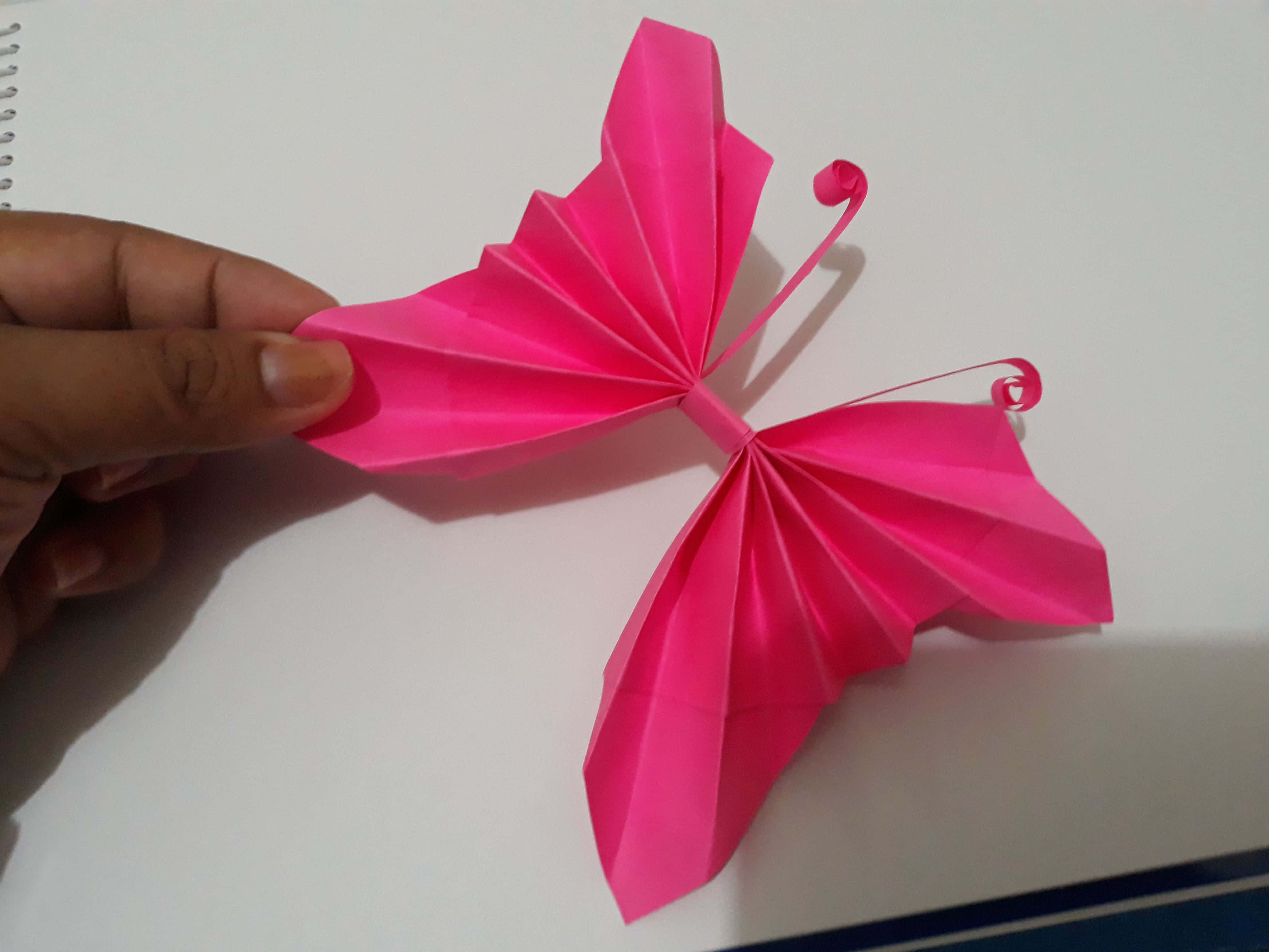 Butterfly Origami 桌布 壁紙 — 一隻手拿著一隻帶有捲曲觸角的鮮豔粉紅色摺紙蝴蝶，背景是純白色筆記本。