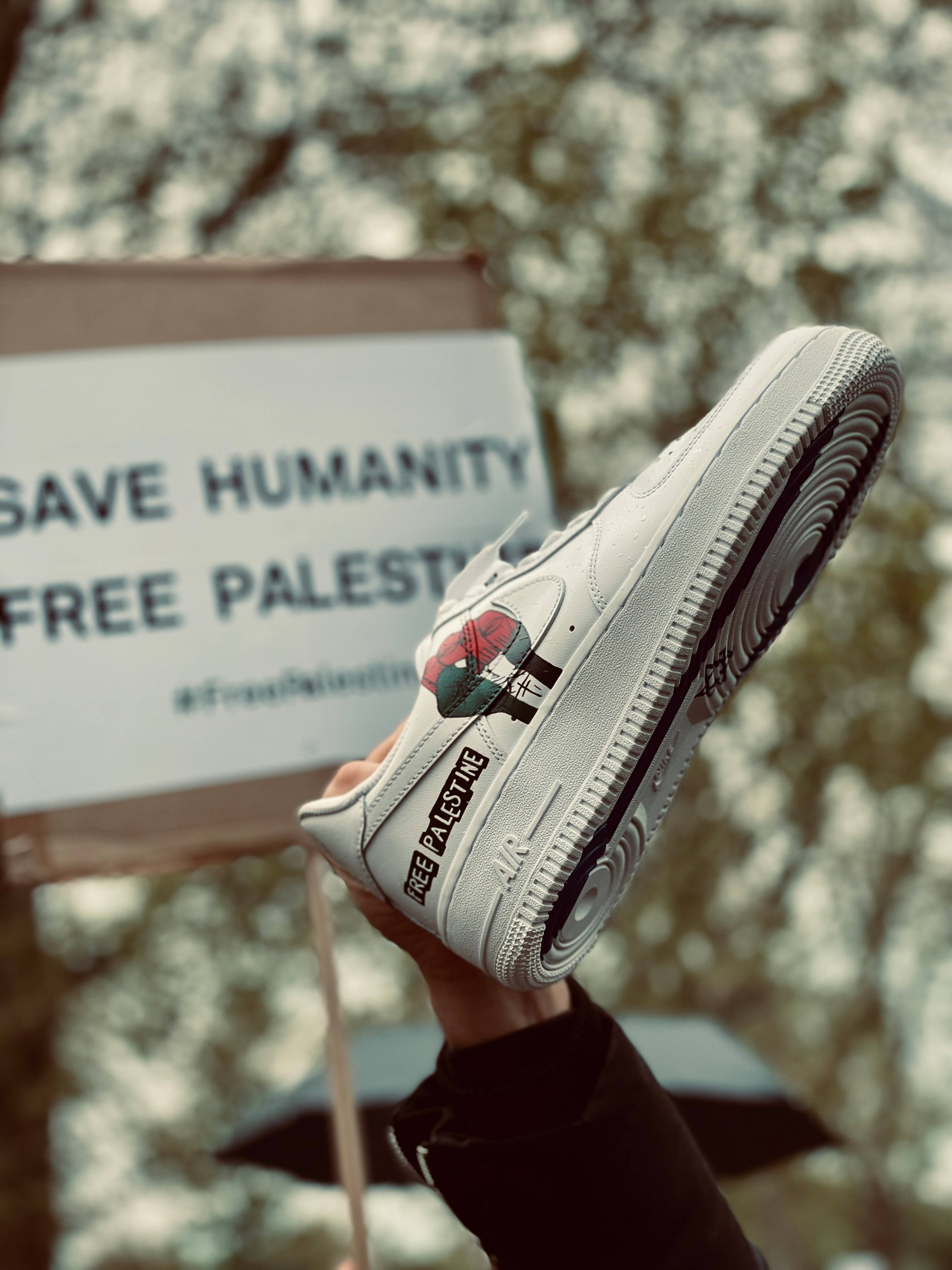 iPad Pro 桌布 壁紙 — 一隻手拿著一隻白色運動鞋，上面有 'FREE PALESTINE' 和一朵玫瑰補丁，位於抗議標語牌上方，標語牌寫著 'SAVE HUMANITY FREE PALESTINE'。