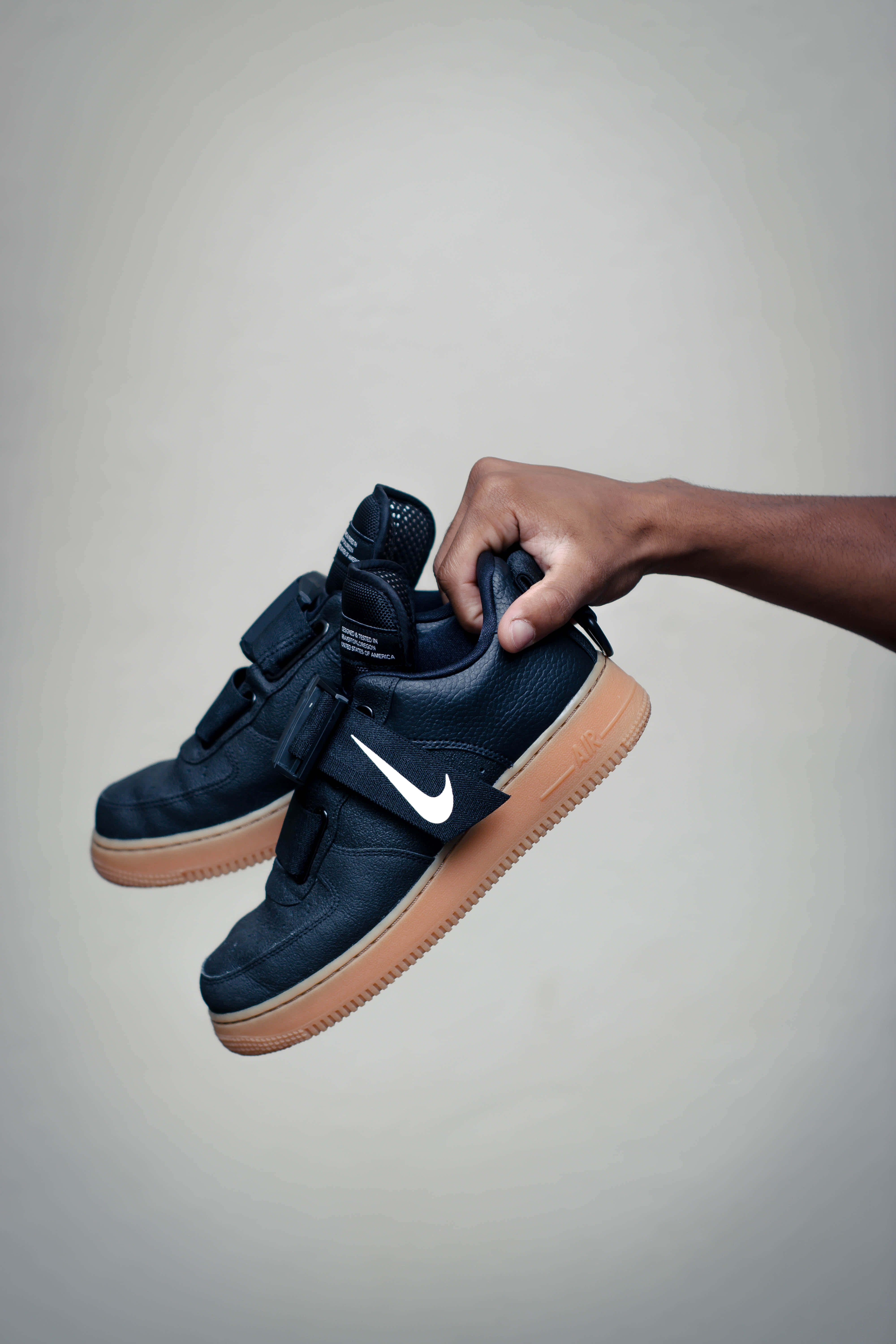 Black Air Force 桌布 壁紙 — 一隻手拿著一雙深藍色Nike運動鞋，帶有白色勾勾和棕褐色鞋底，背景為中性灰色。