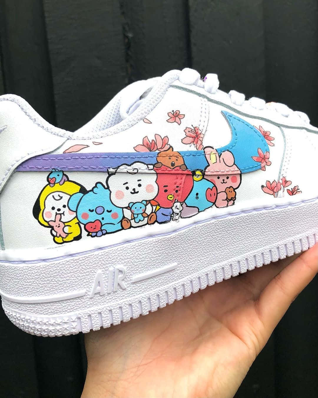 Nike Air Force 桌布 壁紙 — 一隻手拿著一雙白色Nike Air運動鞋，鞋上裝飾著色彩繽紛的BT21卡通角色和粉紅色櫻花，背景為深色木材。