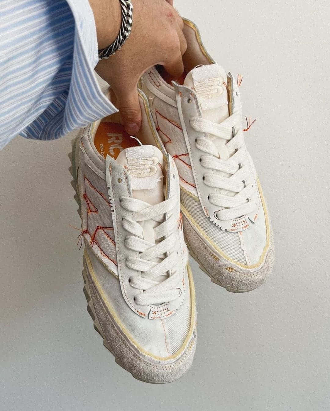 New Balance 桌布 壁紙 — 一隻手拿著一雙白色運動鞋，帶有紅色縫線和磨損的鞋底，靠著素色牆壁。