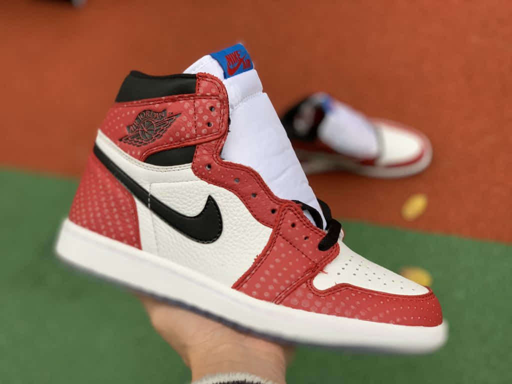 桌布 壁紙 — 一隻手拿著一雙紅白黑配色的Jordan 1運動鞋，鞋面帶有穿孔紋理，背景是模糊的網球場。