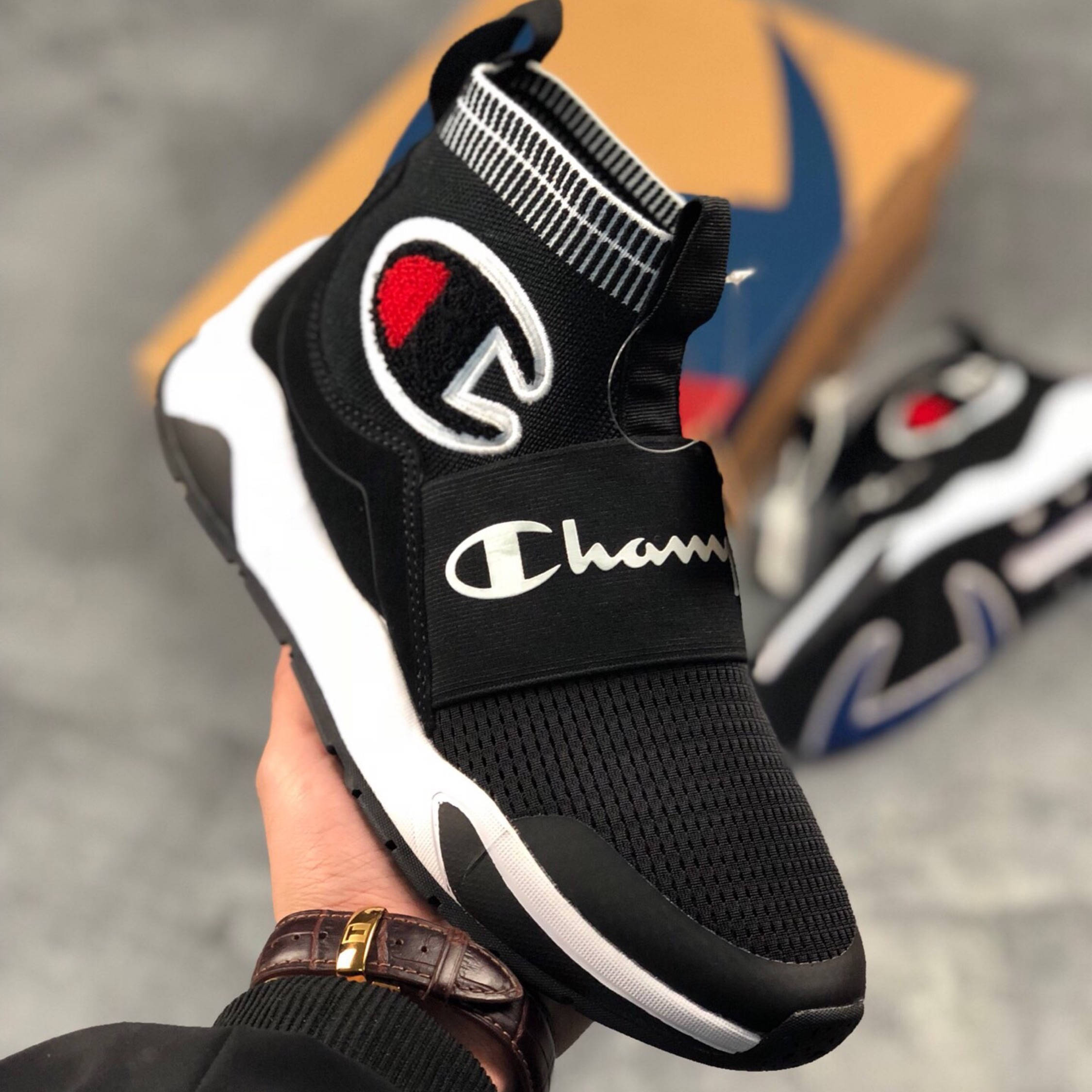 Champion 桌布 壁紙 — 一隻手拿著一雙黑色Champion襪套運動鞋，白色鞋底和紅色標誌，背景是模糊的鞋盒。