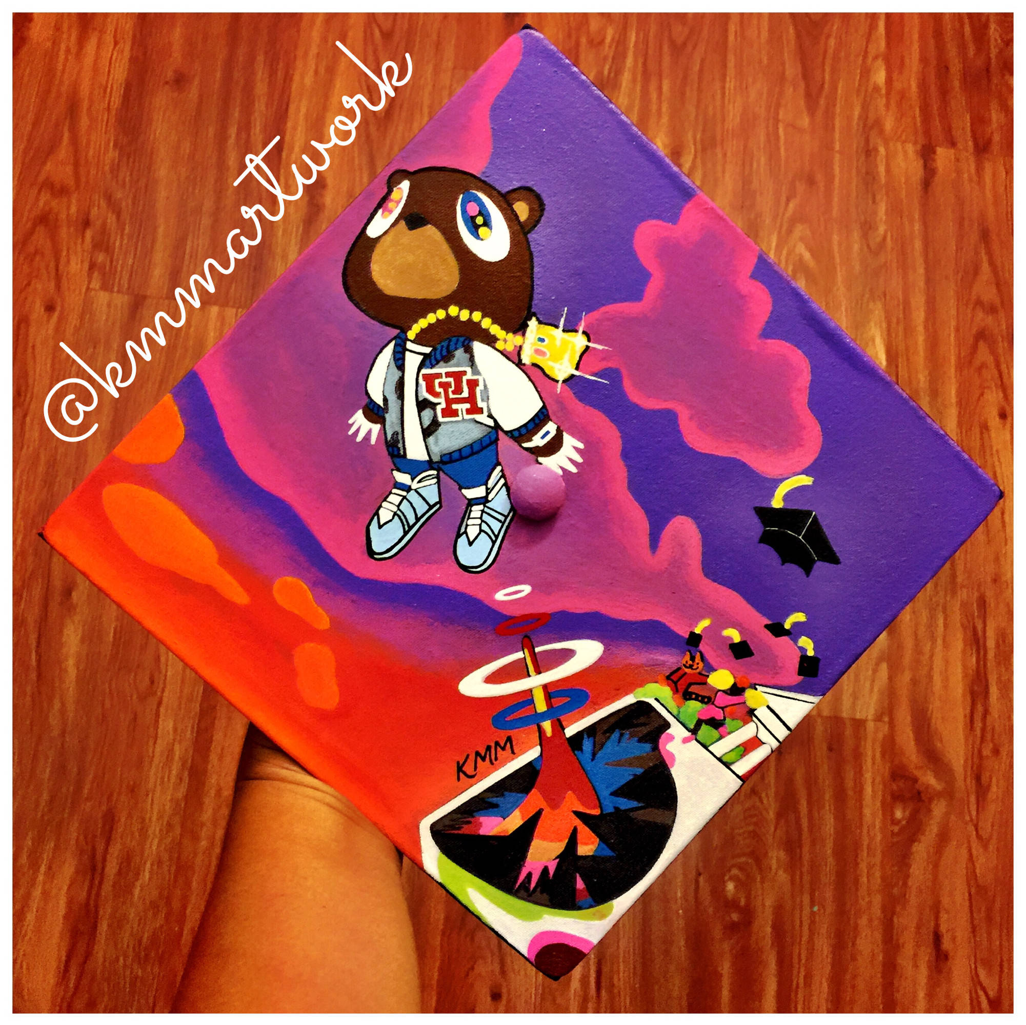 Kanye West Album Cover 桌布 壁紙 — 一隻手拿著一頂畢業帽，上面畫著一隻穿著連帽衫和運動鞋的卡通熊，背景是充滿活力的紫色和橙色。