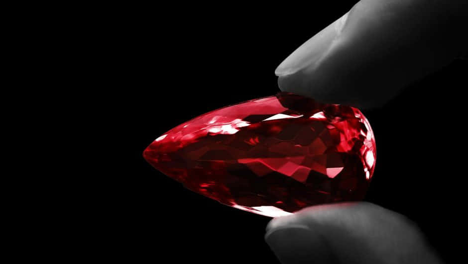 Red Diamond 桌布 壁紙 — 一隻手拿著一顆鮮紅色的梨形寶石，襯托在純黑背景上，突顯其璀璨切面和深邃色彩。