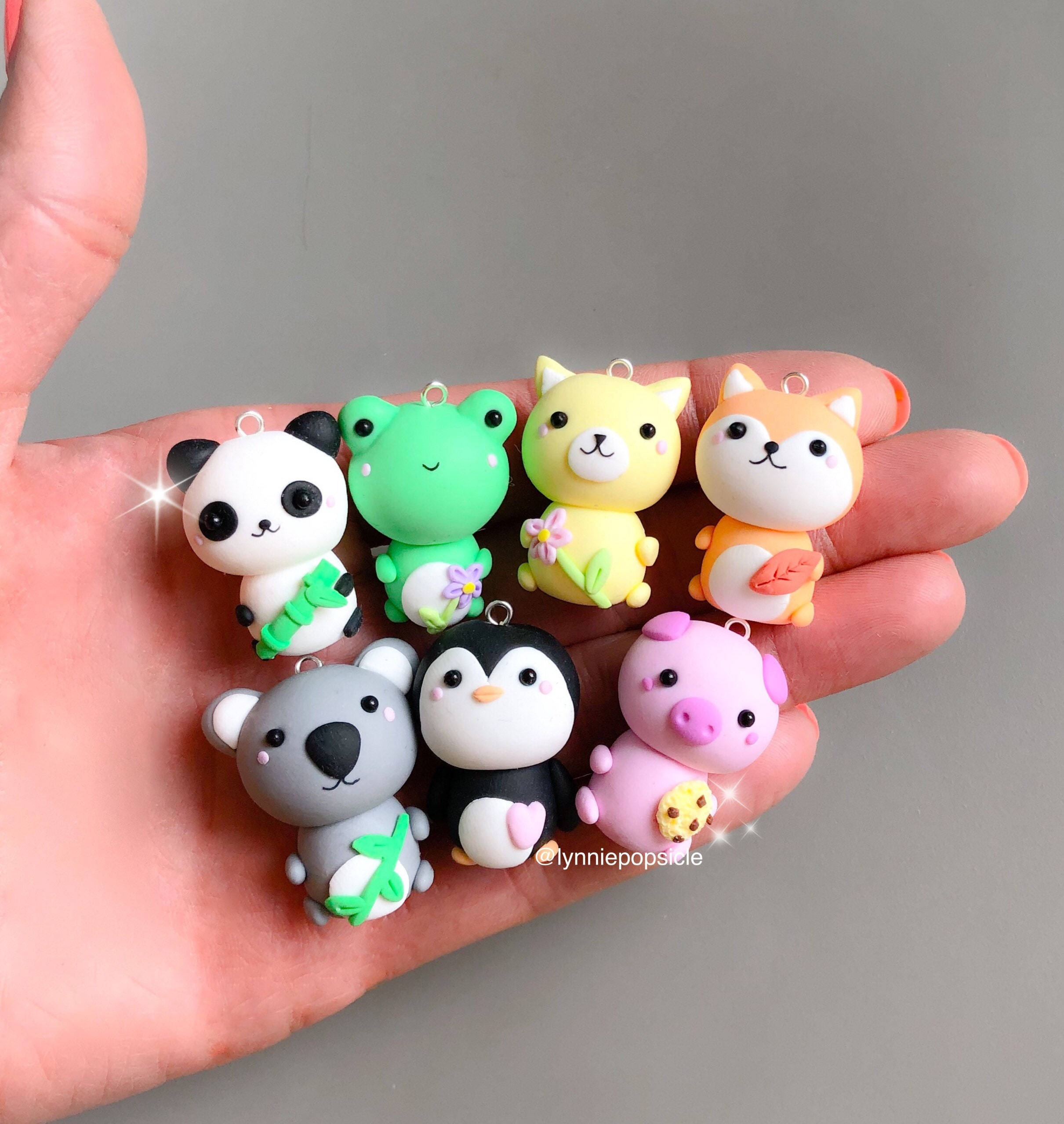 Kawaii Cute Animals 桌布 壁紙 — 一隻手拿著七個色彩繽紛的黏土動物吊飾，包括熊貓、青蛙、貓、狐狸、無尾熊、企鵝和豬，每個都有可愛的配件。