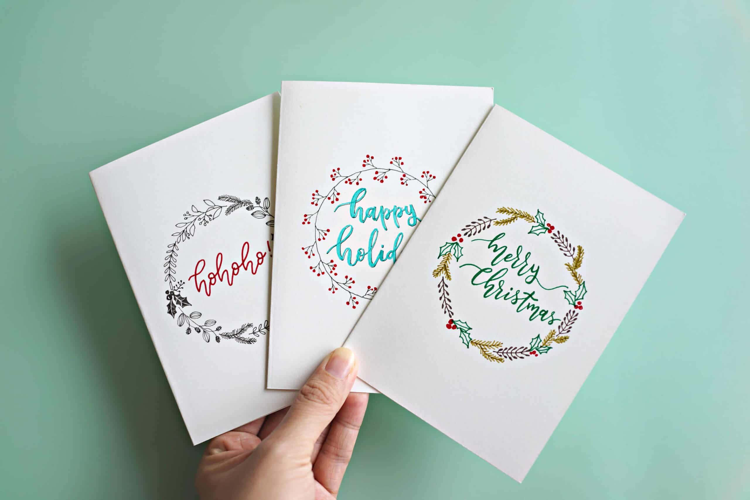 Christmas Card 桌布 壁紙 — 一隻手拿著三張帶有花環設計的節日賀卡，背景為薄荷綠色。