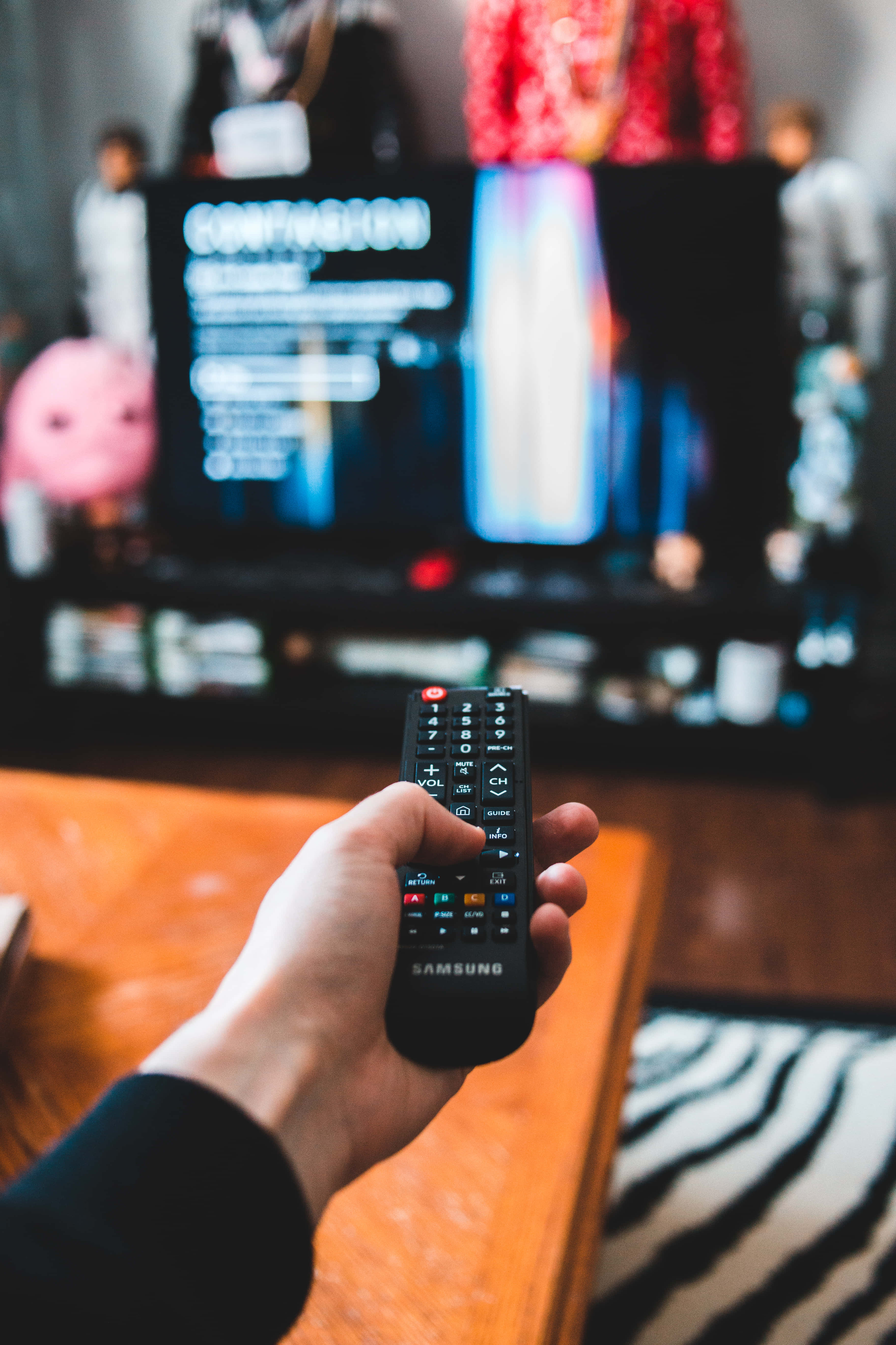 watching tv 桌布 壁紙 — 一隻手拿著三星遙控器，指向一個顯示選單的電視，位於一個有斑馬地毯和絨毛玩具的舒適客廳中。
