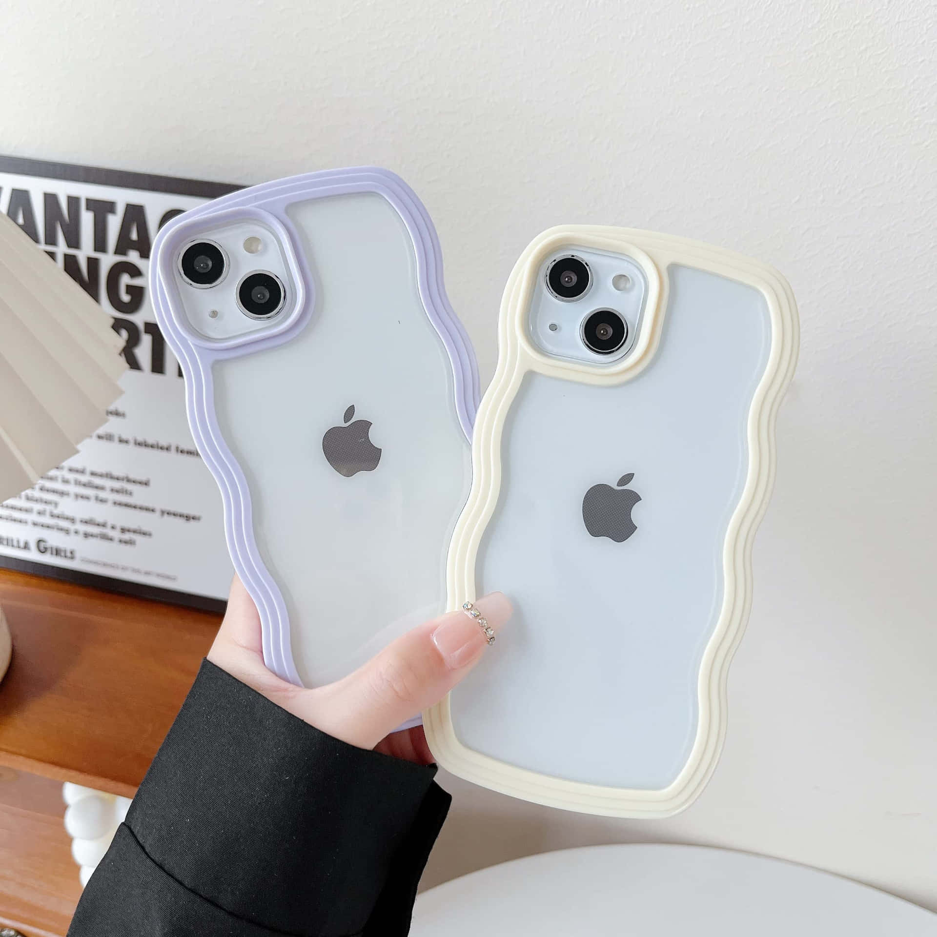 iPhone 美學 桌布 壁紙 — 一隻手拿著兩個波浪邊半透明手機殼，一個薰衣草色一個奶油色，背景是白色牆壁，上面有海報和木架。